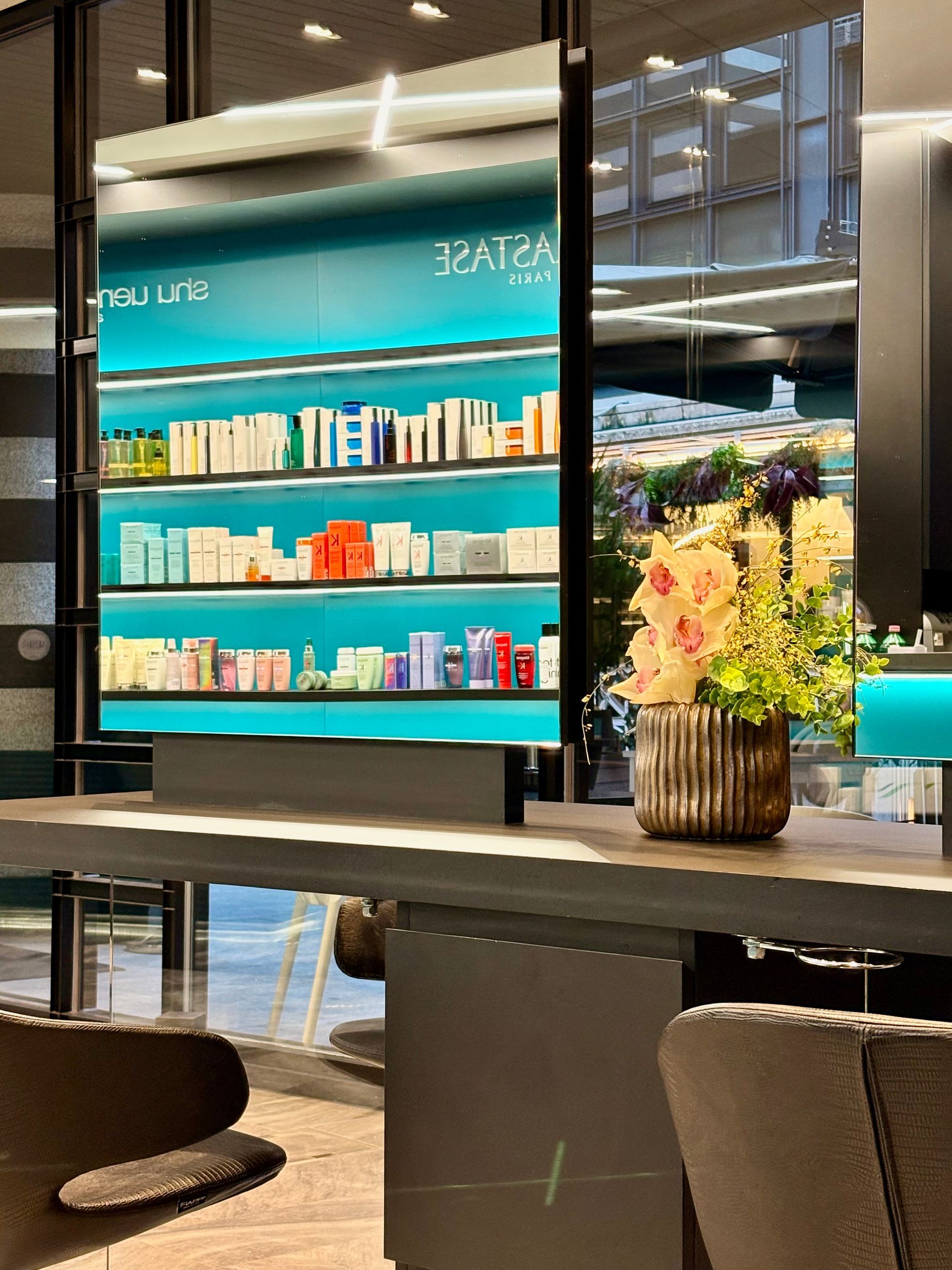 Una postazione da salone con uno scaffale a specchio contenente vari prodotti per la cura dei capelli e un vaso decorativo di orchidee.