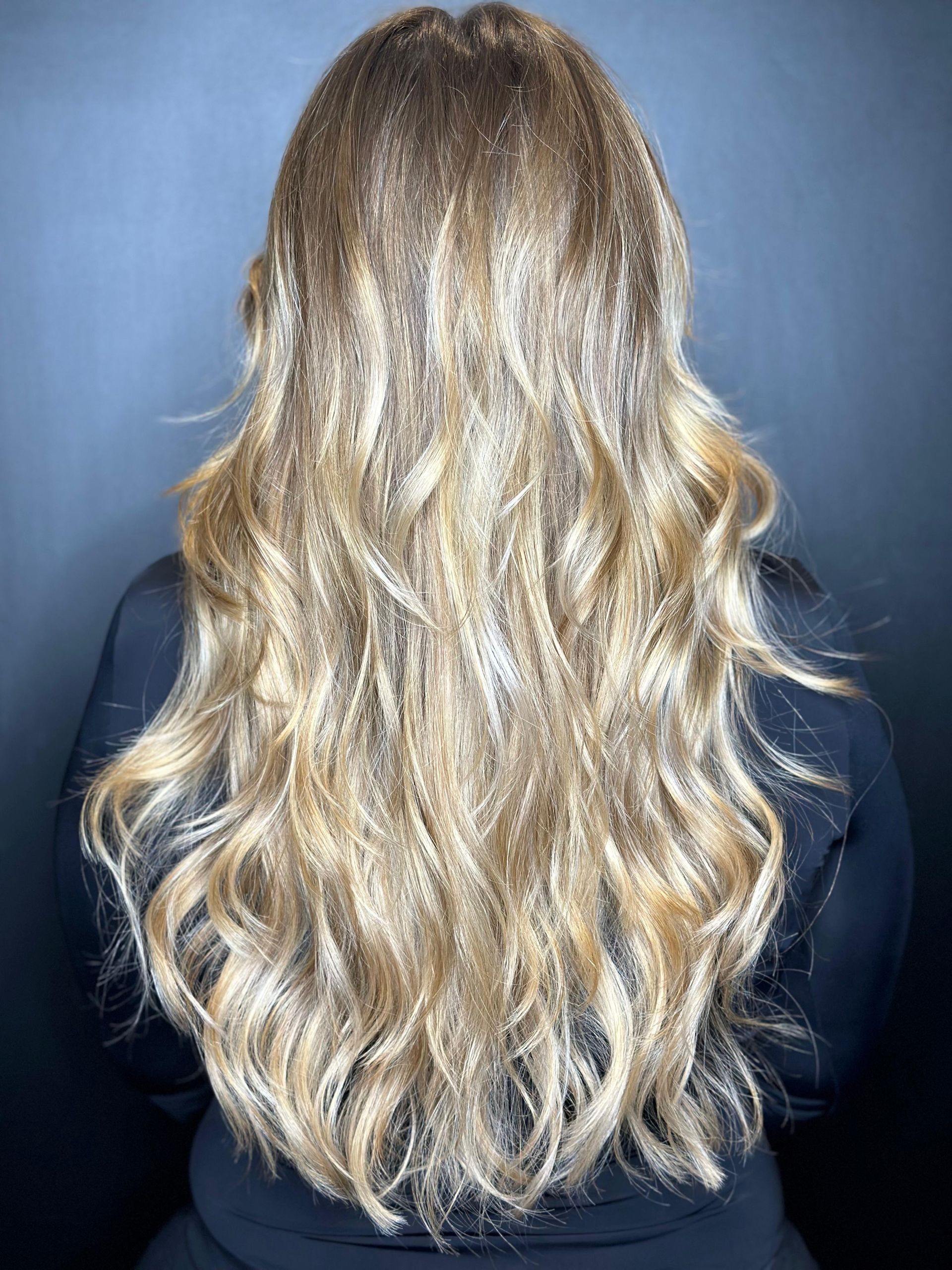 Capelli lunghi e ondulati con riflessi biondi e radici più scure, visti di spalle su uno sfondo grigio scuro. MAGAMO Lugano