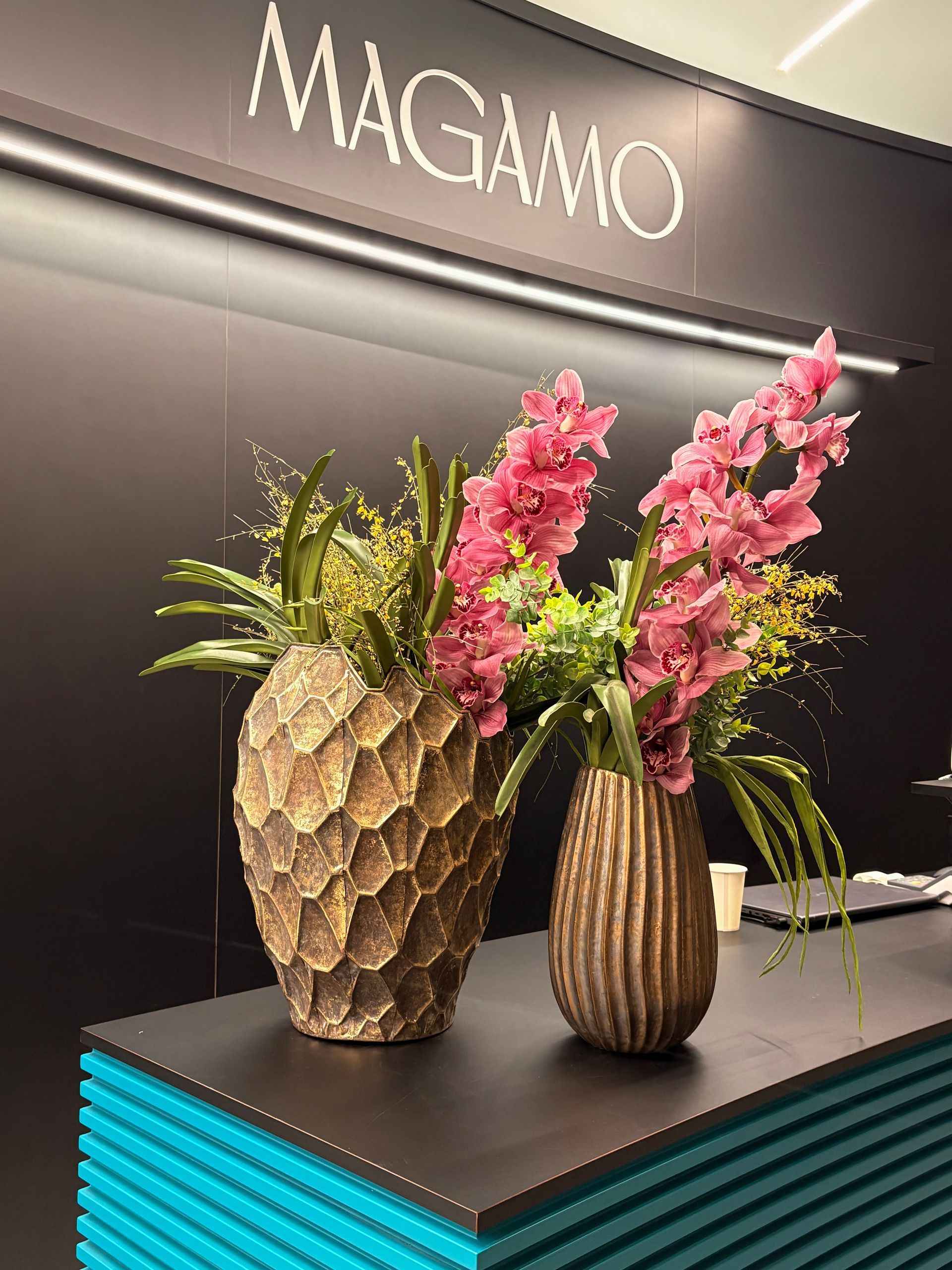 Due vasi dalla superficie ruvida, contenenti orchidee rosa e fogliame verde, sono appoggiati su un ripiano scuro davanti a un cartello con la scritta MAGAMO.
