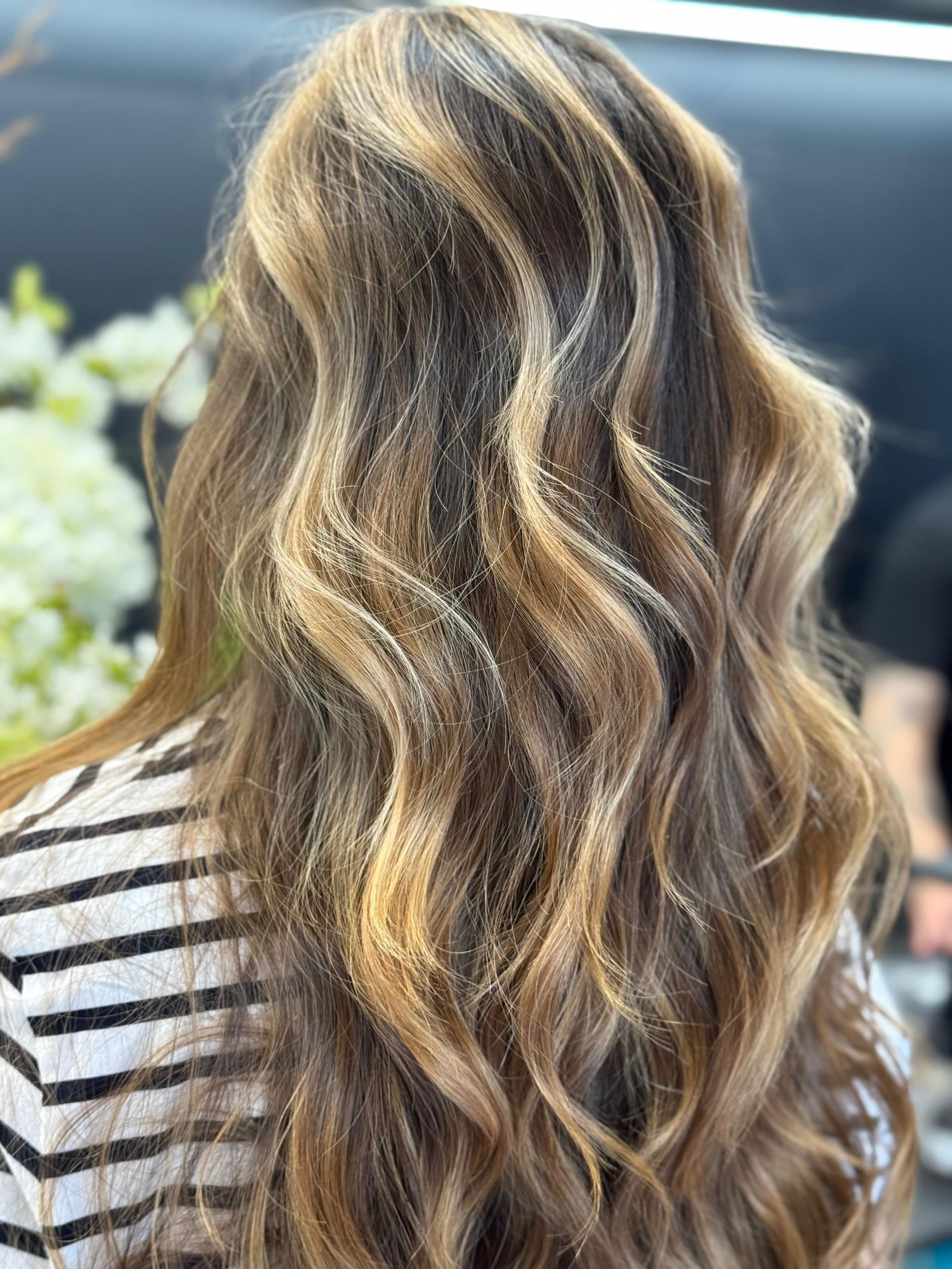 Vista posteriore di lunghi capelli ondulati con riflessi biondi, acconciati in morbidi ricci, indossati sopra una camicia a righe. MAGAMO Lugano