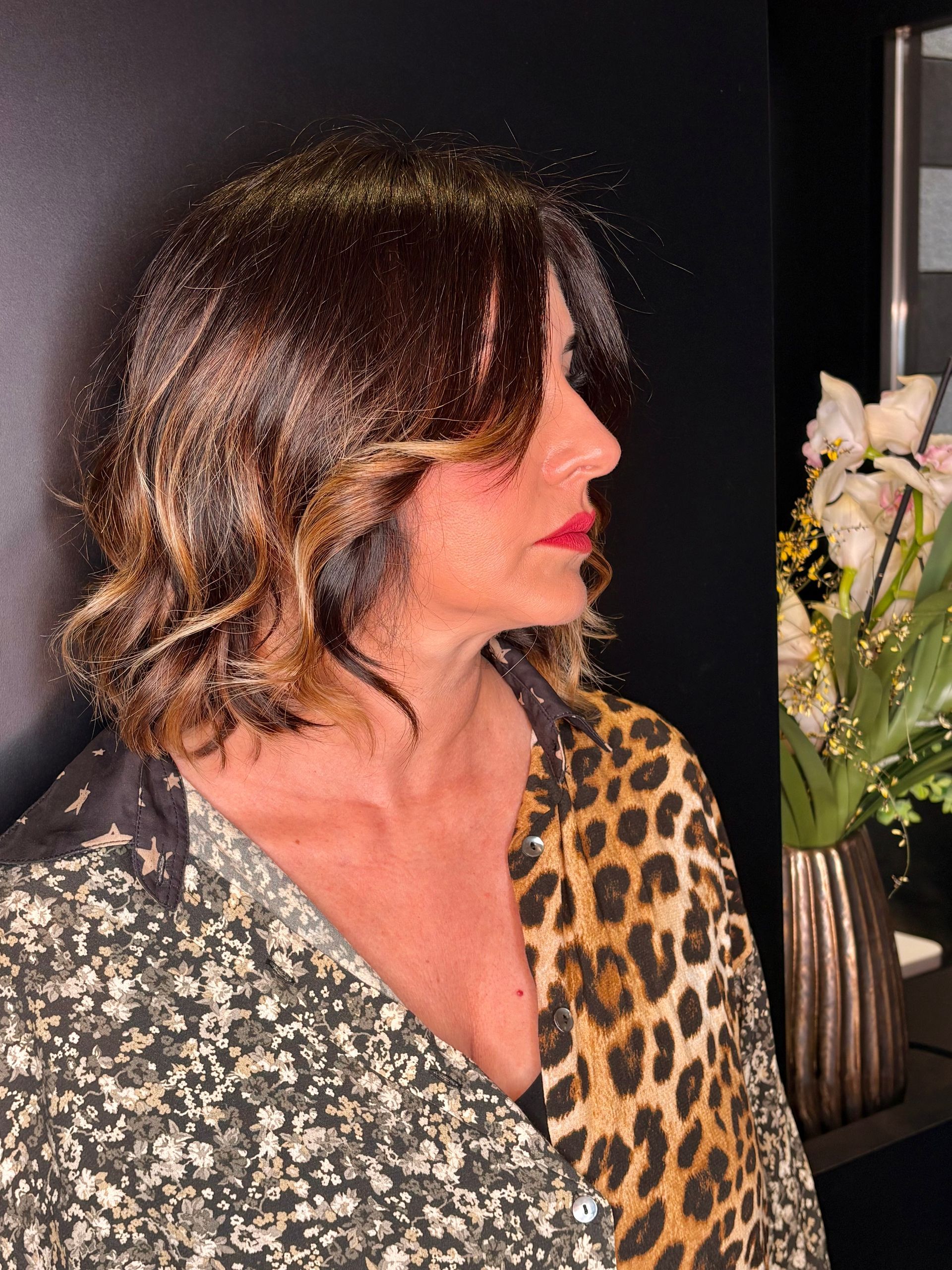 Una persona con i capelli mossi lunghi fino alle spalle e il rossetto rosso indossa una camicia a fantasia mista, in posa vicino a un vaso di fiori.
