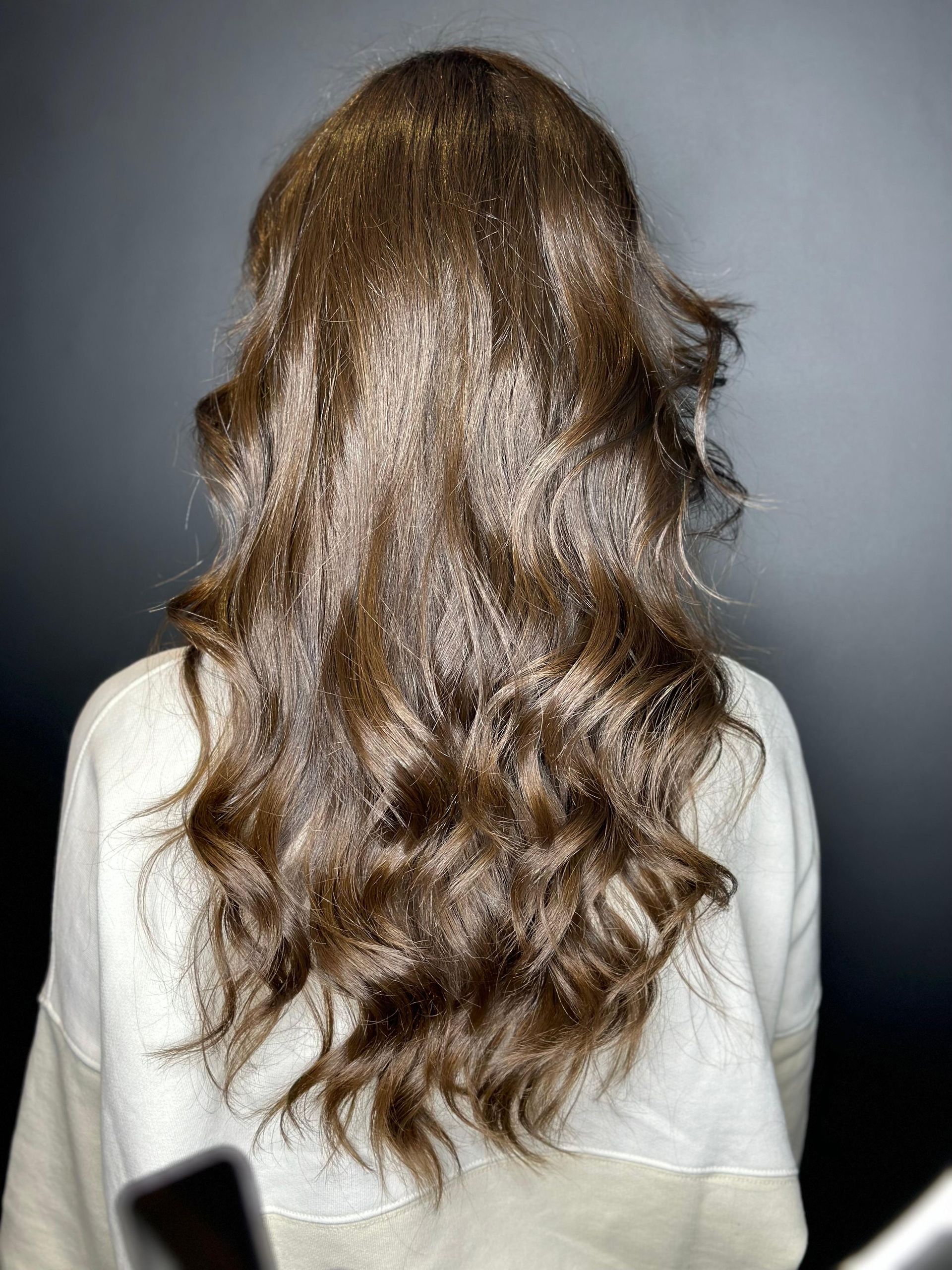 Una persona con capelli castani ondulati, lunghi fino alle spalle, vista di spalle, indossa un maglione color crema su uno sfondo grigio. MAGAMO Lugano