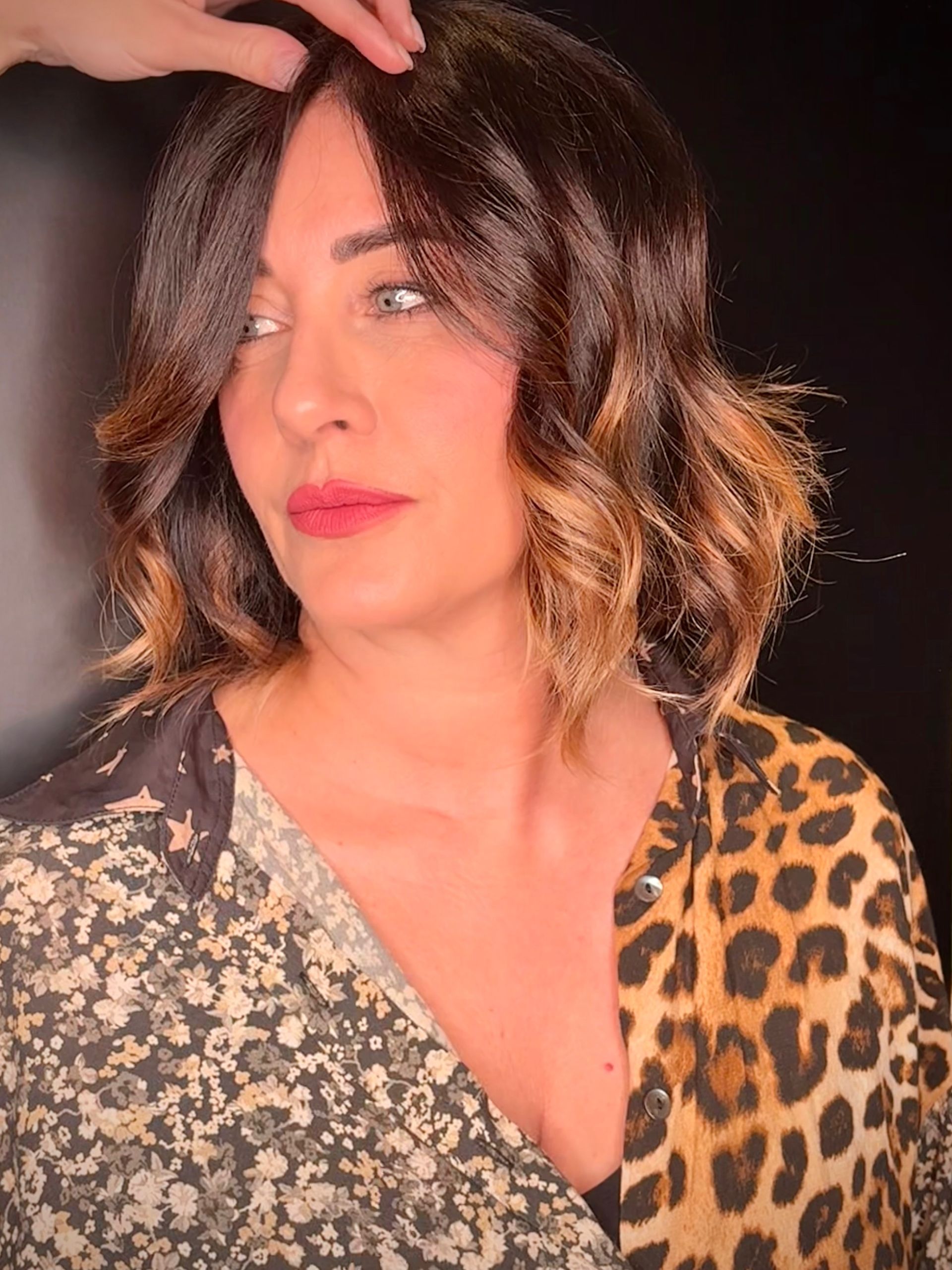 Primo piano di una persona con capelli mossi, di colore dal castano scuro al castano chiaro, e rossetto rosso, che indossa una maglietta a fantasia, vista di profilo.