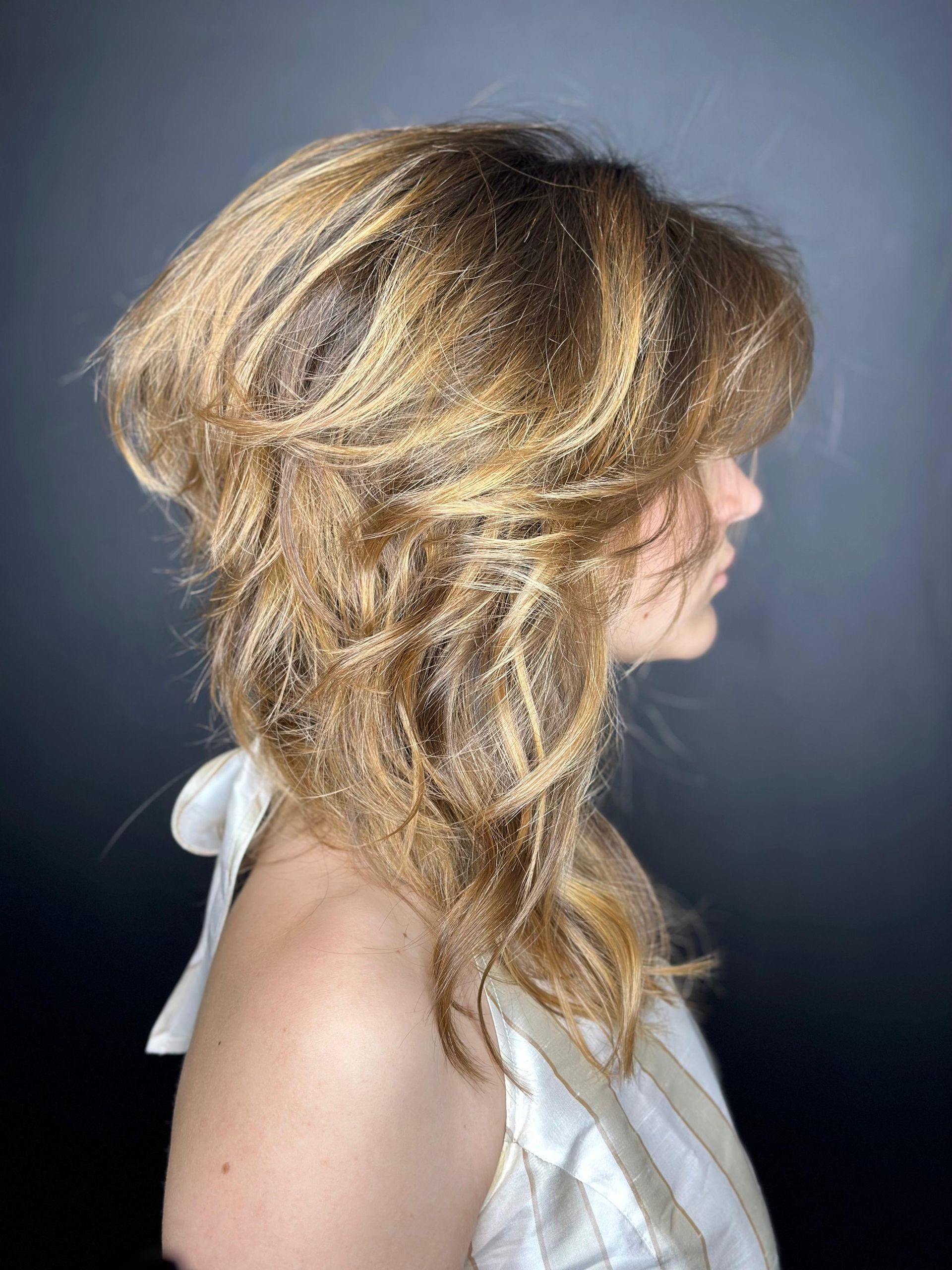 Profilo laterale di una persona con capelli biondo dorato, mossi e ricci, acconciati in morbide onde, su uno sfondo grigio scuro uniforme. MAGAMO Lugano