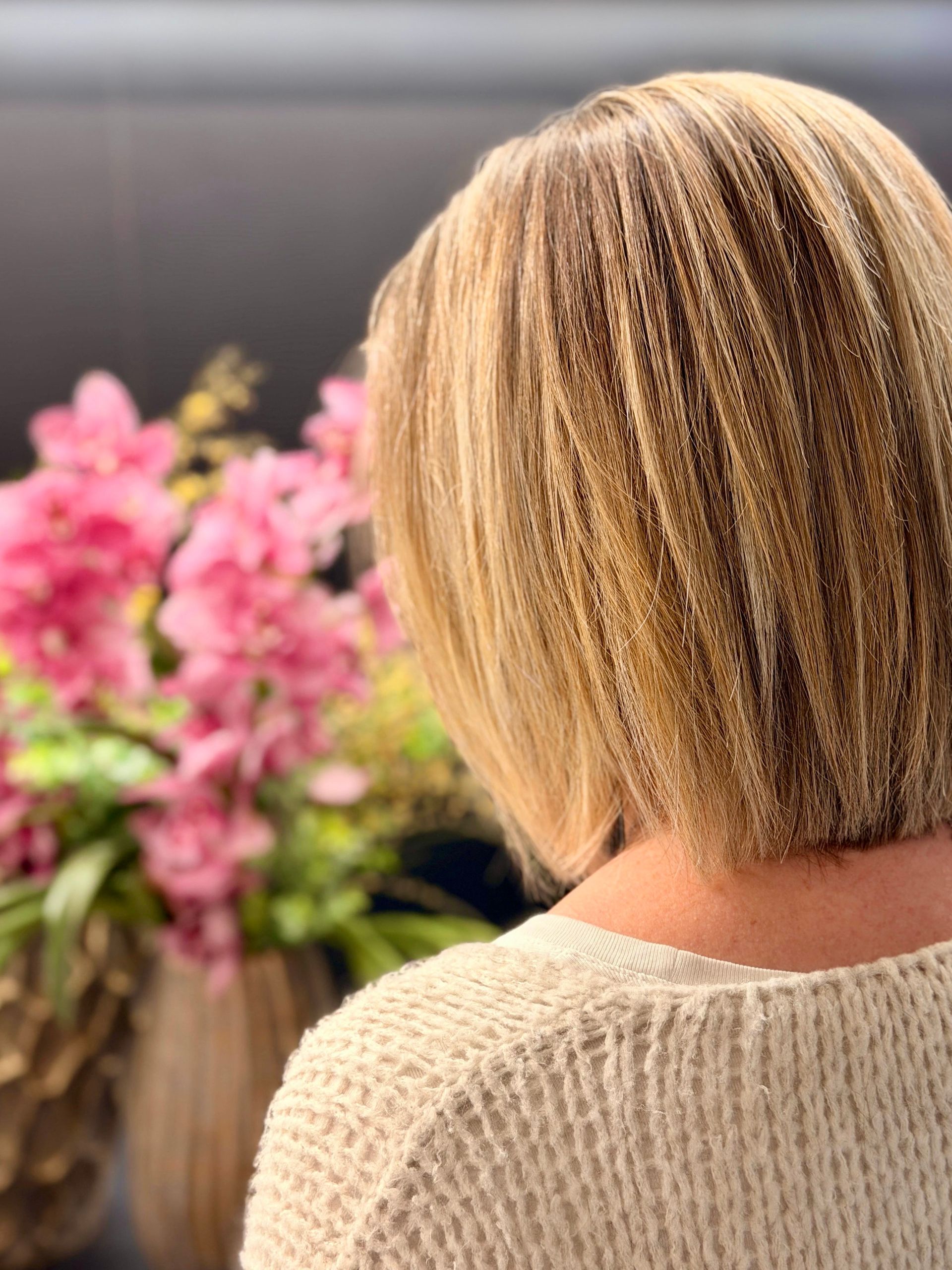 Vista posteriore di una persona con capelli biondi, ondulati e di lunghezza caschetto, che indossa un maglione color crema dalla trama particolare, accanto a orchidee rosa.