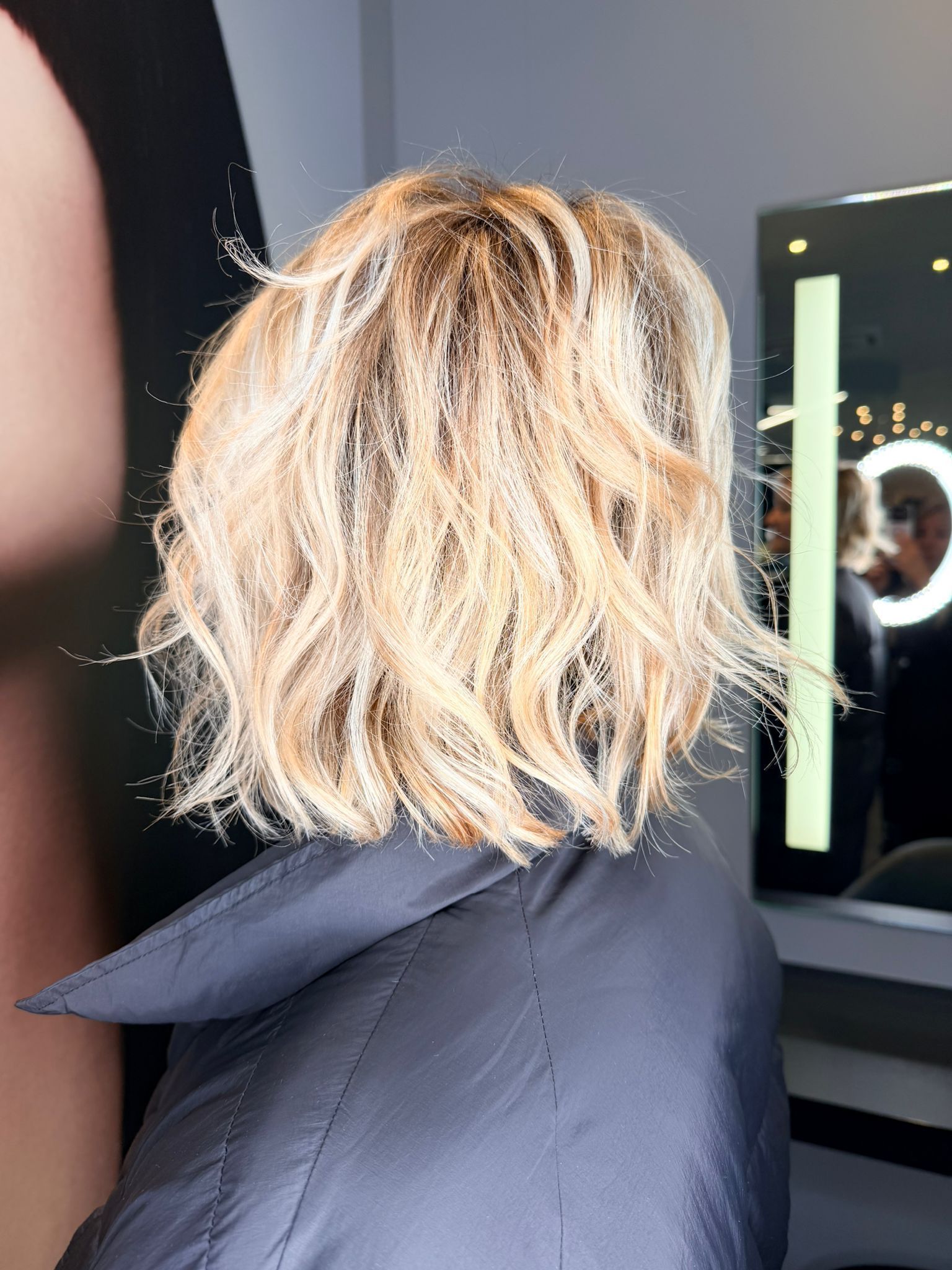 Vista posteriore di capelli biondi, mossi e lunghi fino alle spalle, in un salone di bellezza, con una luce ad anello visibile nello specchio sullo sfondo. MAGAMO Lugano