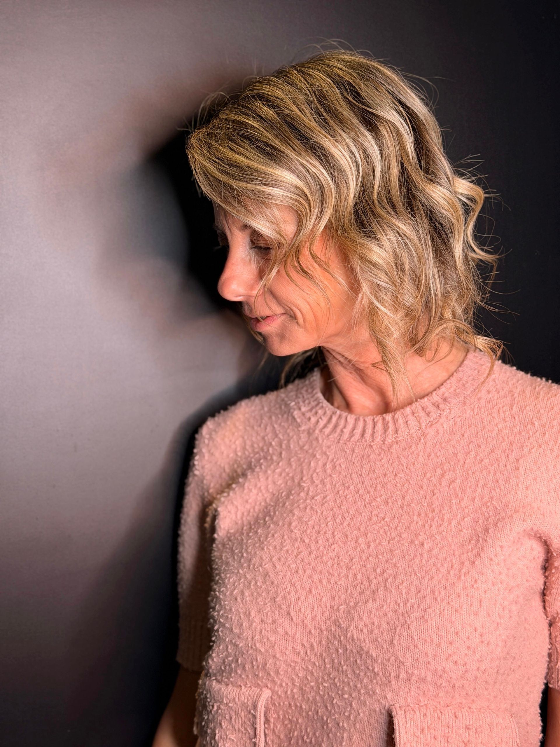 Una persona con i capelli biondi ondulati indossa un maglione di lana rosa su uno sfondo scuro e in ombra.