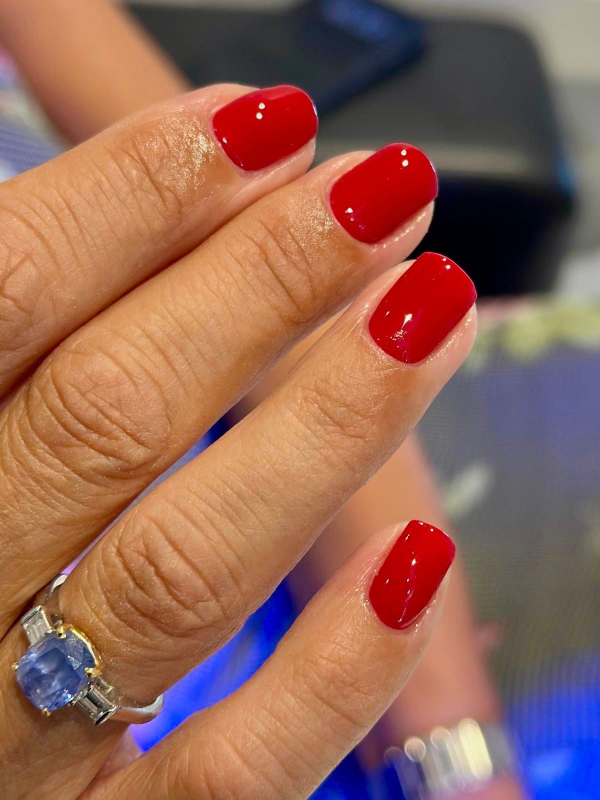Una mano con un anello di pietra preziosa blu e unghie corte, arrotondate e ben curate, di un rosso acceso.