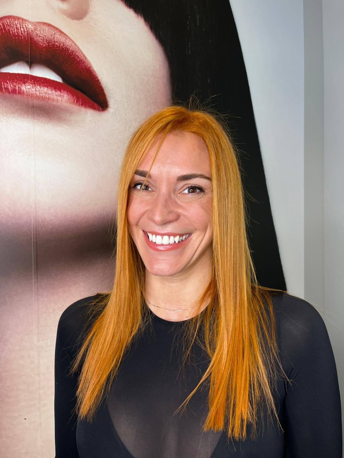Una persona sorridente con lunghi capelli rosso vivo indossa una maglietta nera davanti a un grande ritratto di labbra rosse. MAGAMO Lugano