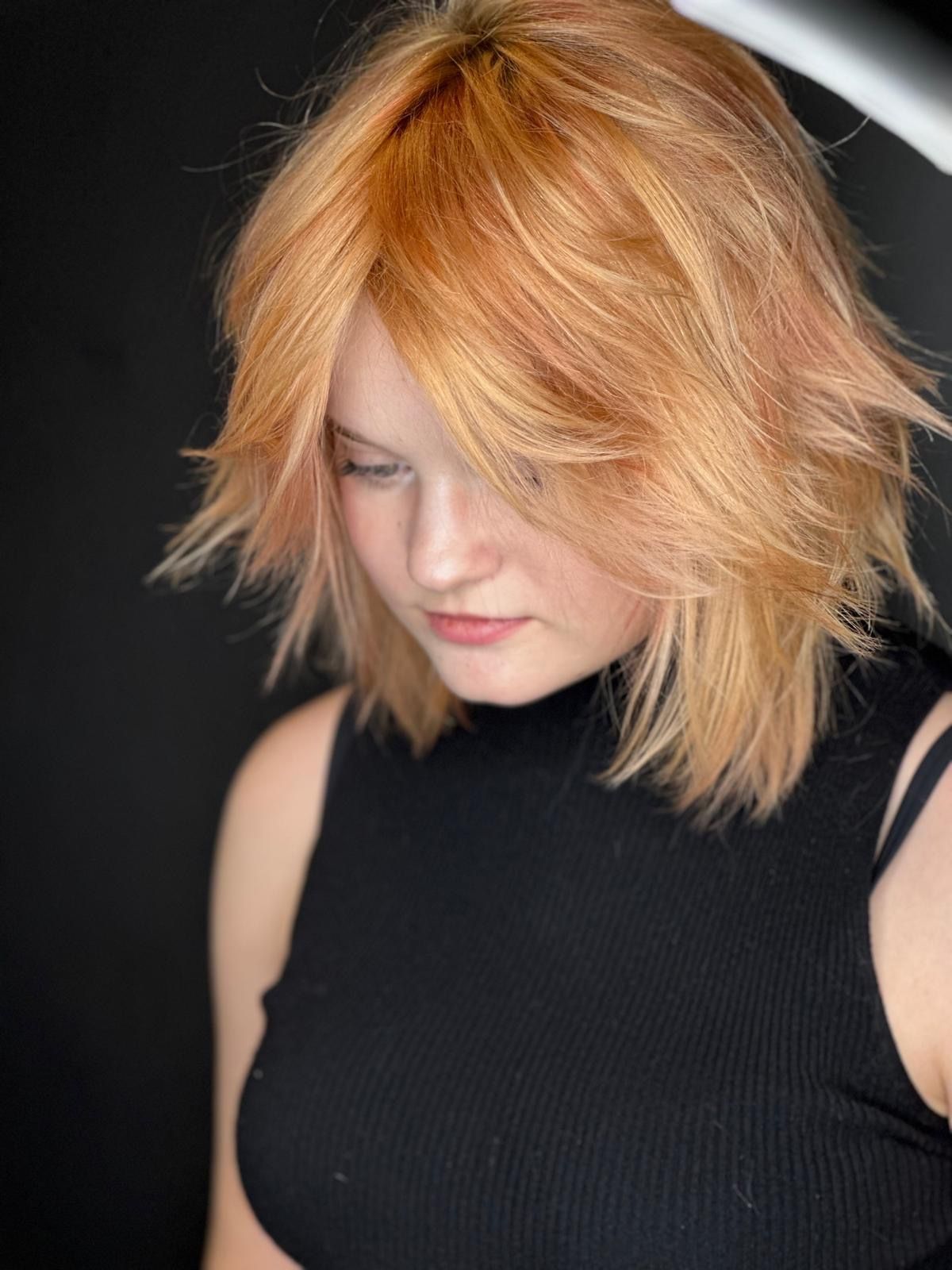 Persona con capelli biondo fragola scalati, lunghi fino alle spalle, che indossa una canotta nera senza maniche e guarda in basso.