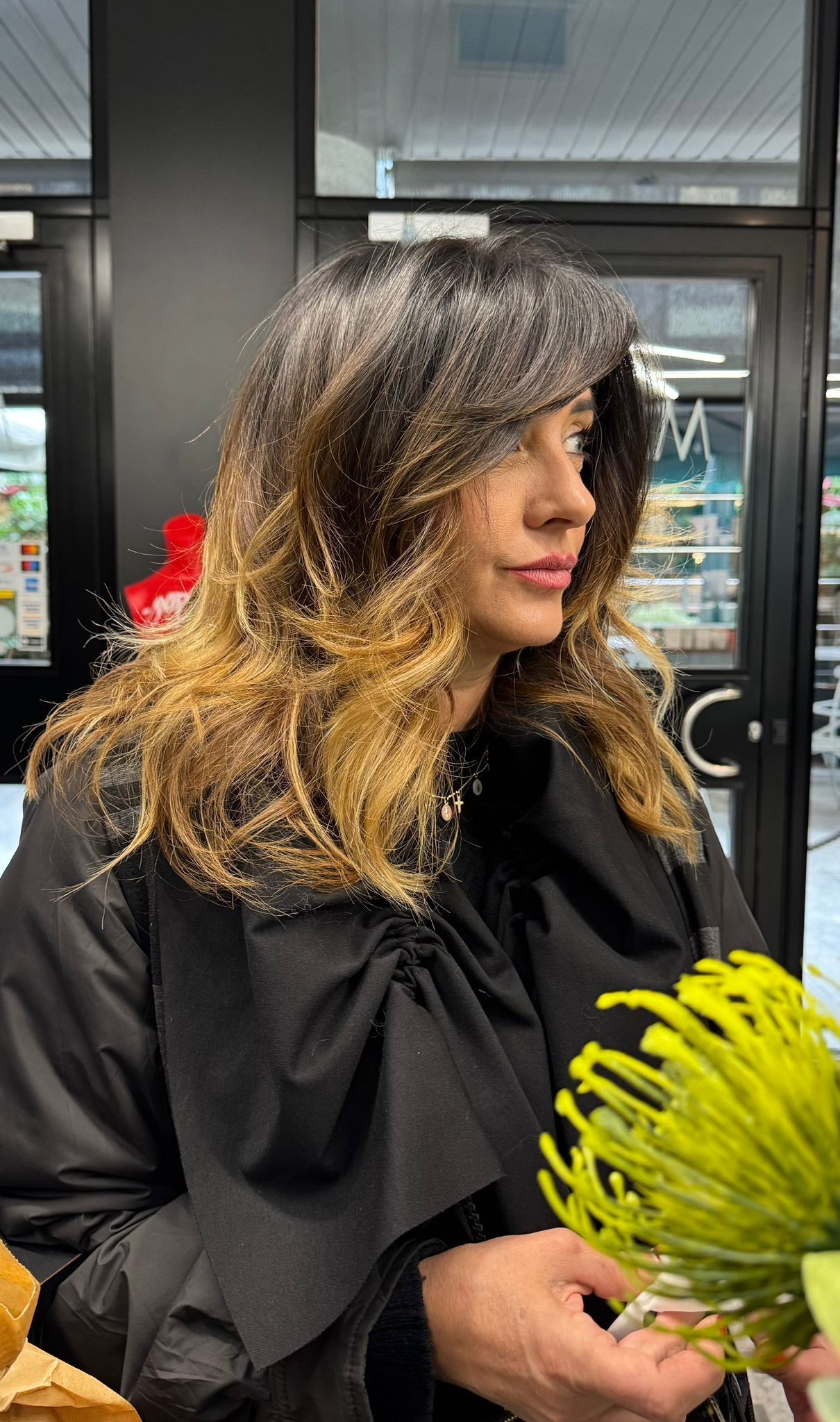 Una persona con i capelli mossi, di colore che sfuma dal scuro al chiaro, tiene in mano un fiore verde brillante davanti alla vetrina di un negozio.