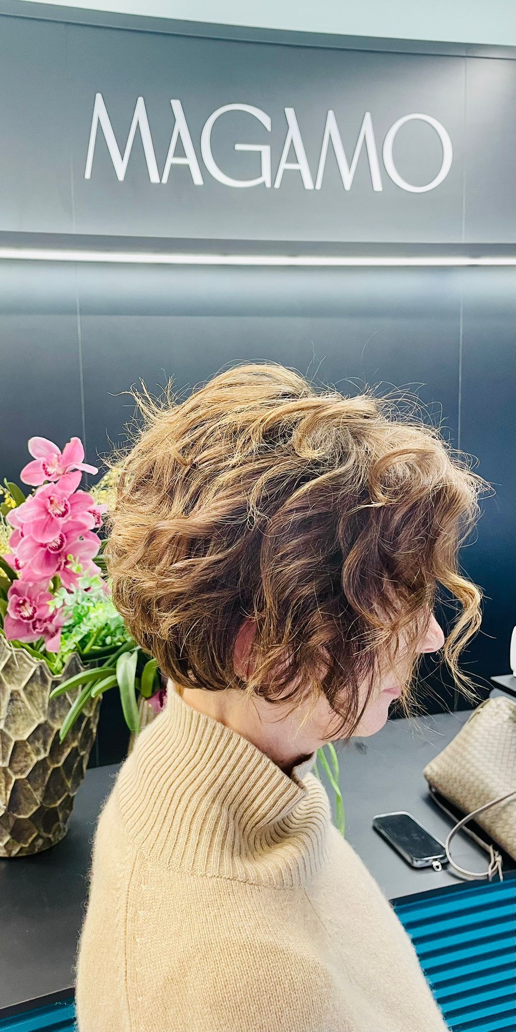 Una persona con i capelli castani ricci, lunghi fino alle spalle, che indossa un maglione color cachi, è in piedi davanti a un cartello con la scritta MAGAMO.