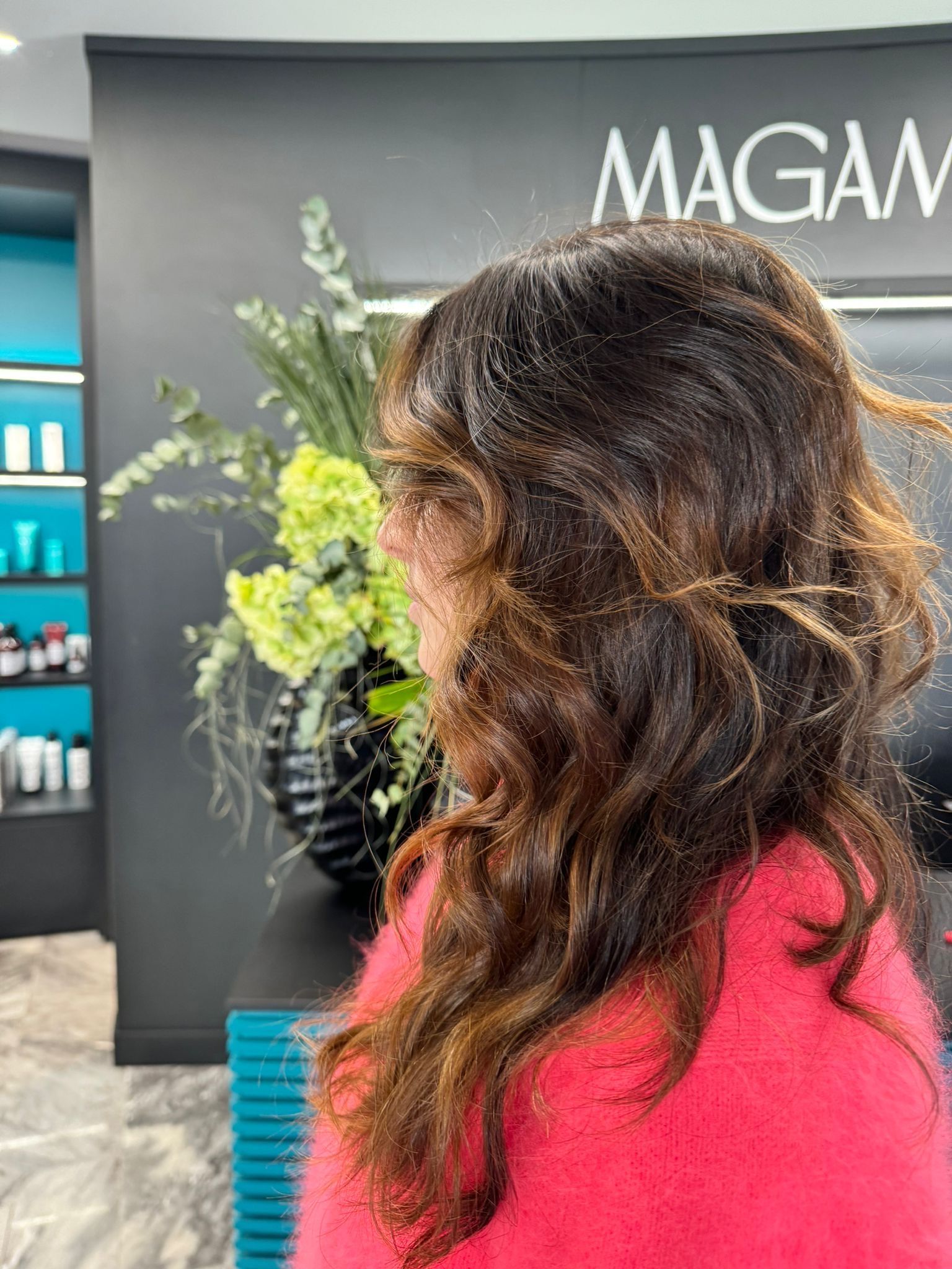 Una persona con i capelli mossi e castani con mèches, che indossa un maglione rosa acceso, è in piedi in un salone di parrucchiere con decorazioni floreali verdi.