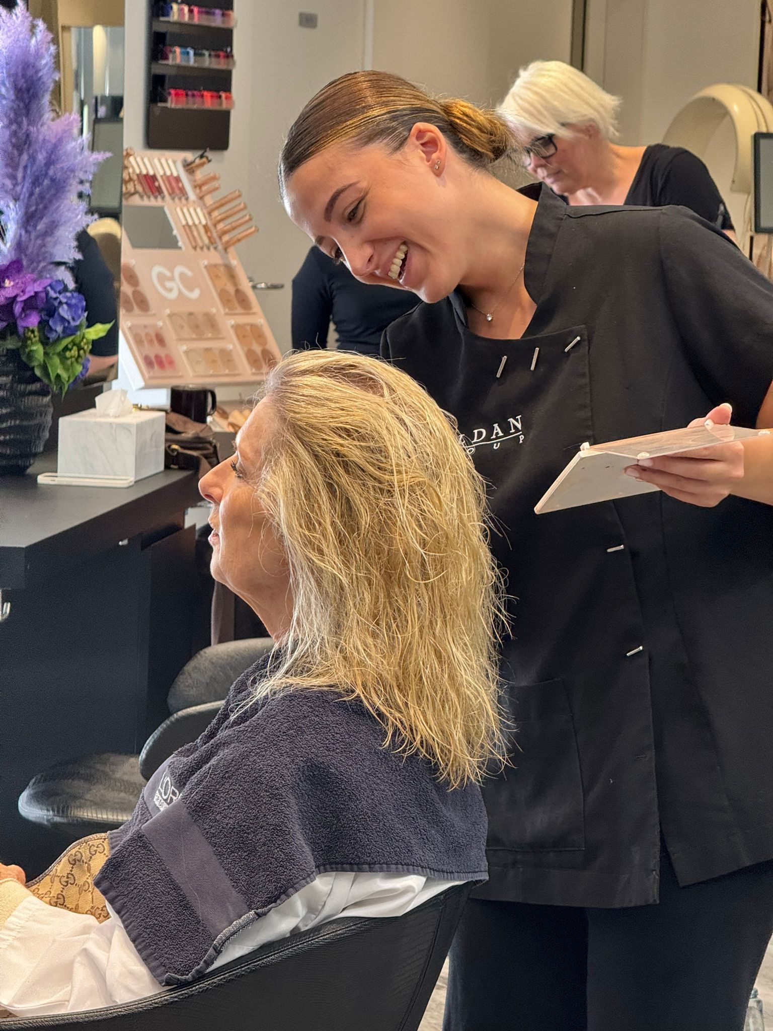 In un salone di parrucchiere, una parrucchiera sorride mentre parla con una cliente dai capelli biondi e ricci.