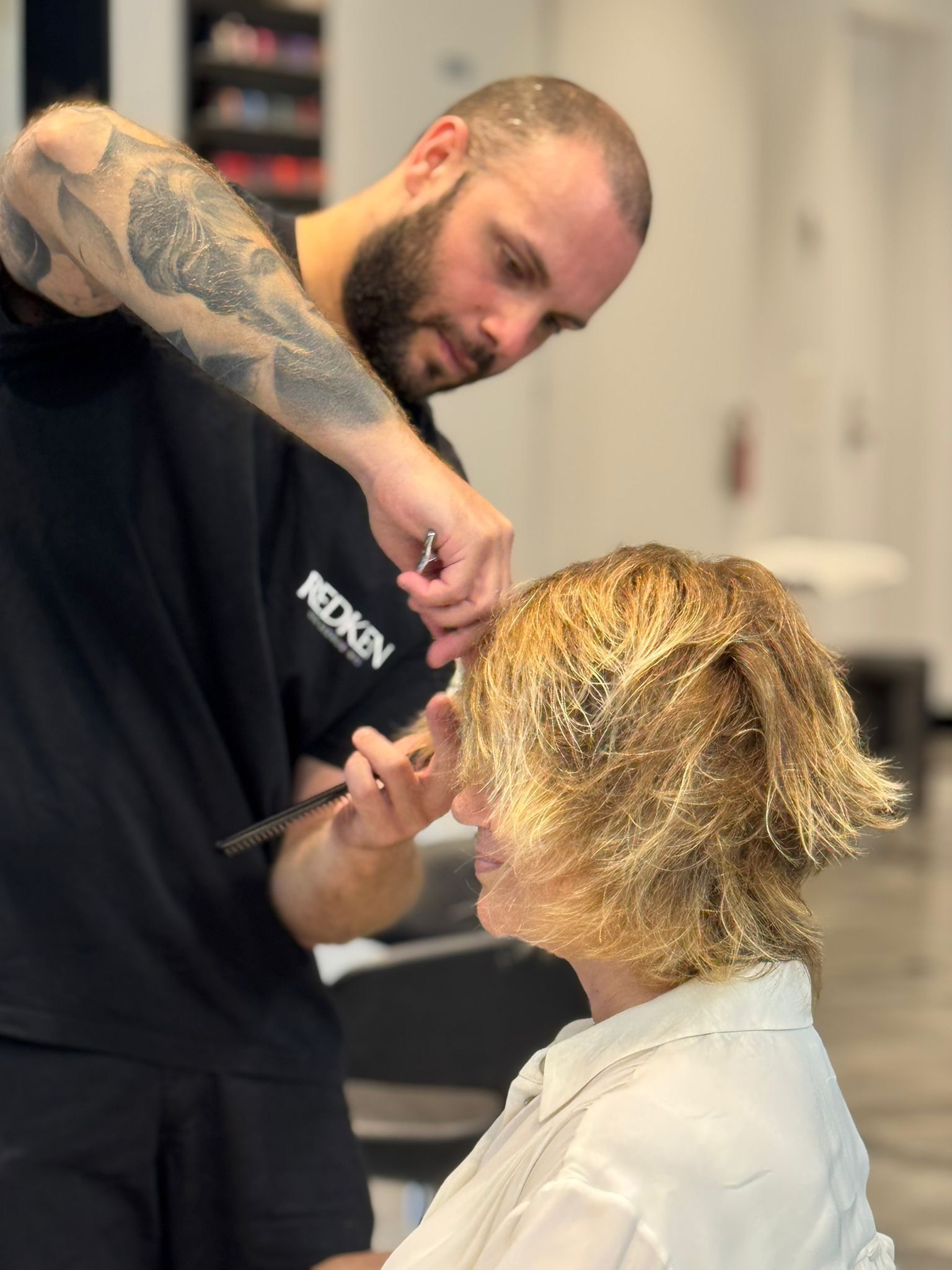 In un salone luminoso, una parrucchiera con un braccio tatuato taglia i capelli biondi di una cliente. MAGAMO Lugano