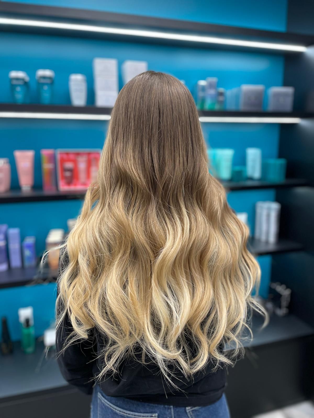 Una persona con lunghi capelli castani ondulati che sfumano verso il biondo alle punte è in piedi davanti agli scaffali di un negozio.