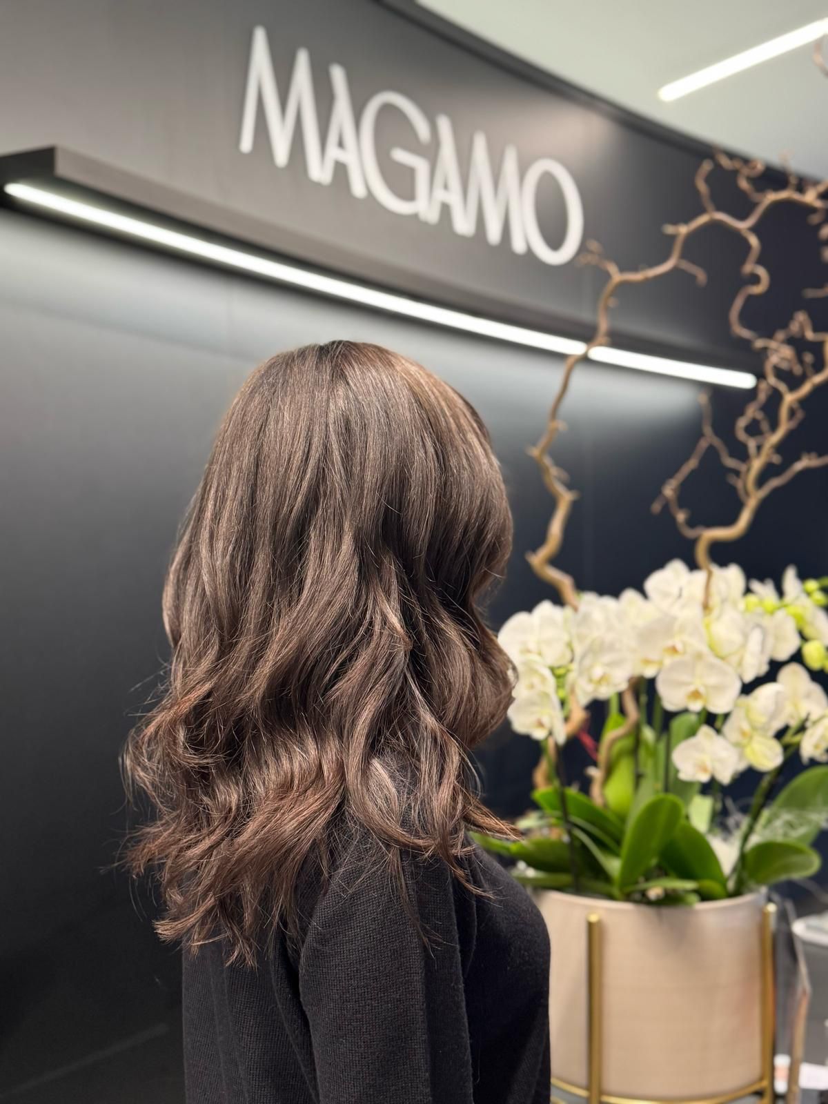 Una persona con i capelli castani ondulati è in piedi davanti a un muro nero con la scritta bianca 