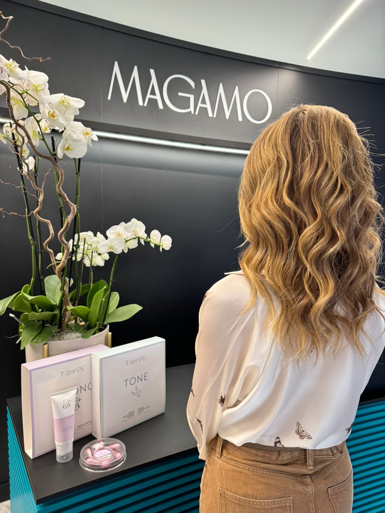 Una persona con lunghi capelli biondi e ondulati si trova davanti a un espositore del marchio Magamo, su cui sono esposte orchidee bianche e prodotti di bellezza.