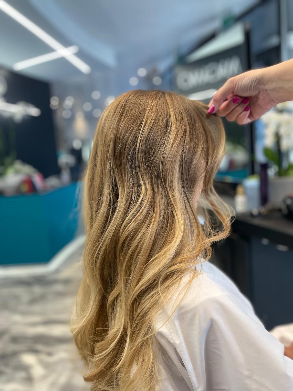 In un salone di bellezza, un parrucchiere tiene in mano una ciocca di capelli lunghi, mossi e biondo dorato.