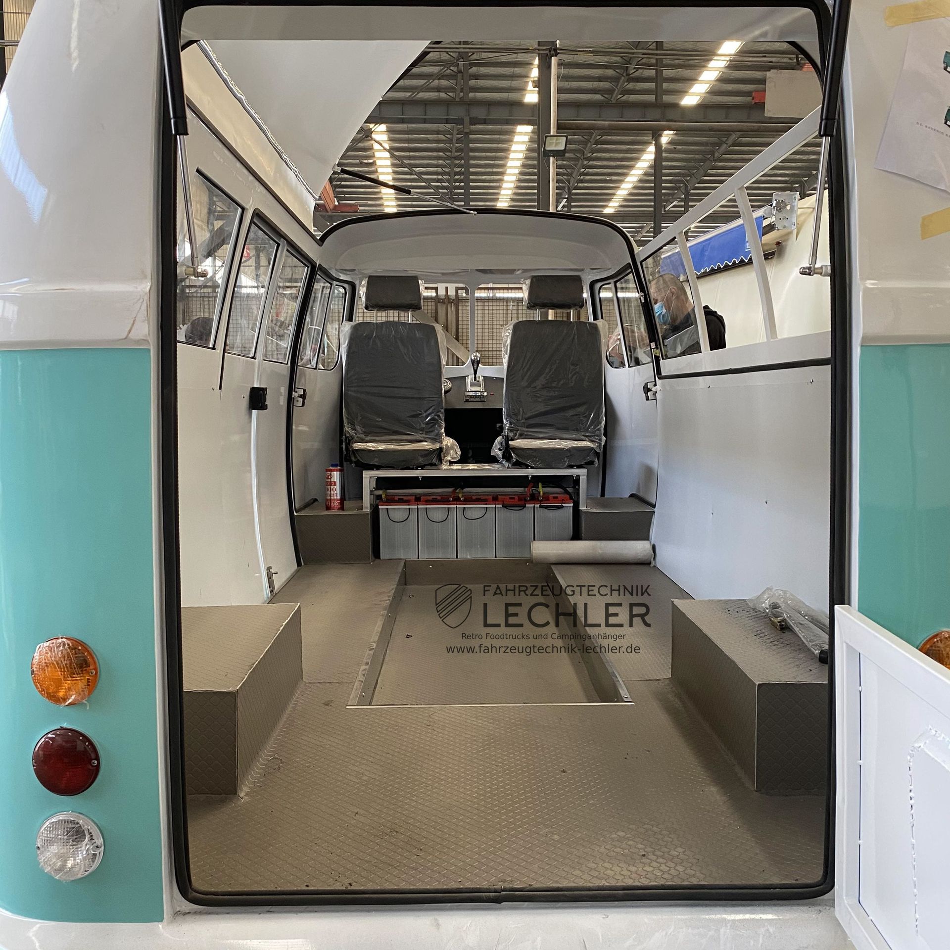 Innenraum eines hellblauen und weißen Lieferwagens, mit Sitzen, Batterien und Stauraum, von hinten gesehen.