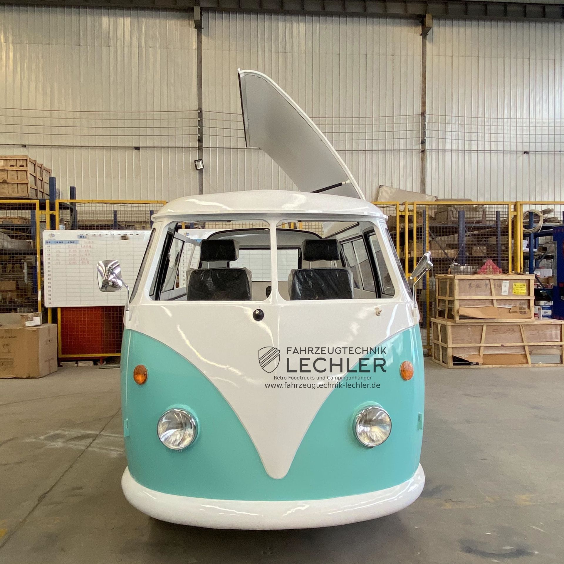 Ein türkisfarbener und weißer VW-Bus mit hochgeklapptem Schiebedach in einer Werksumgebung.