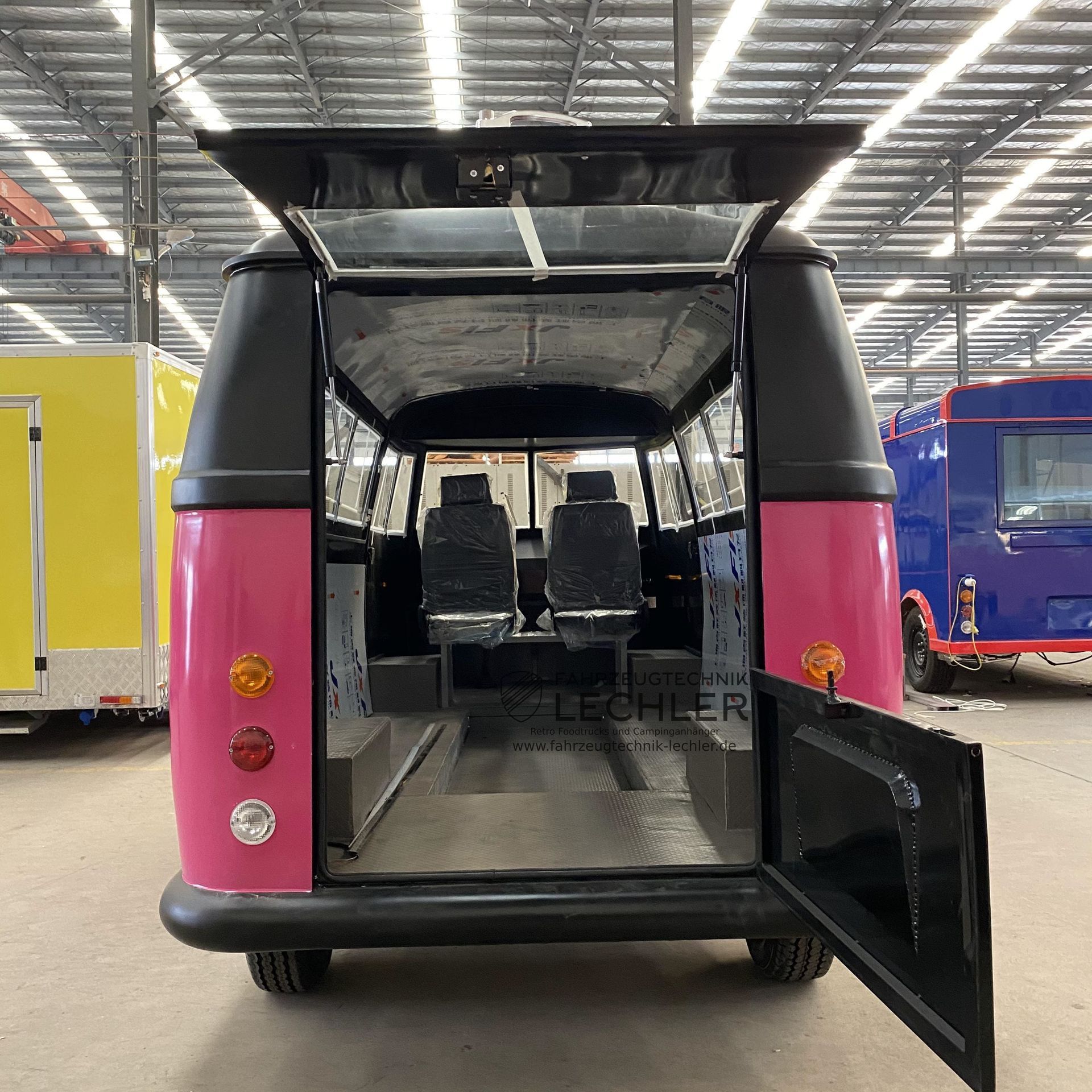 Pinkfarbener und schwarzer Imbisswagen, Rückansicht mit geöffneten Türen, zeigt den Innenraum mit Sitzplätzen, geparkt in einer Halle.