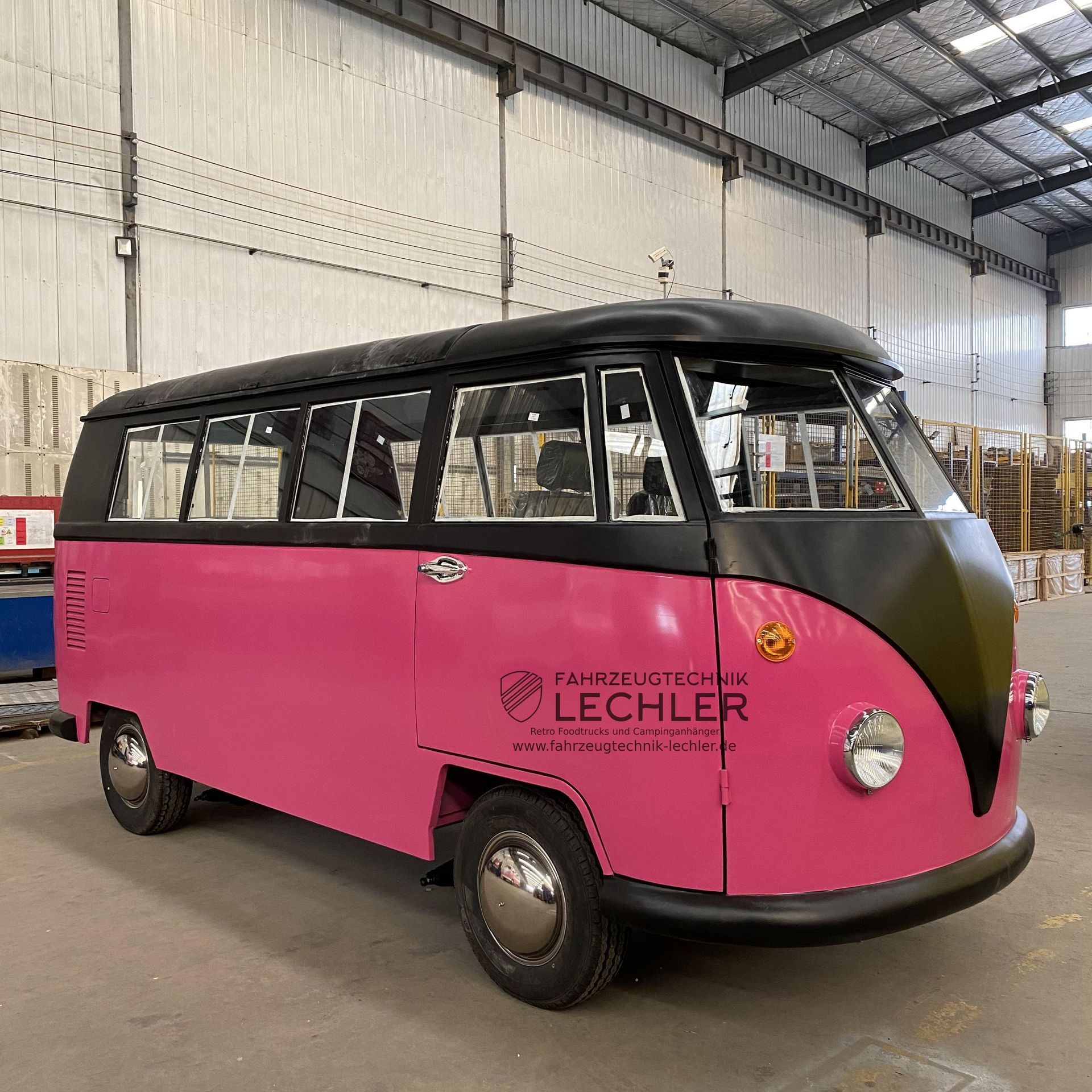 Ein pinkfarbener und schwarzer Volkswagen-Bus in einem Industriegebiet, mit Logo an der Seite.