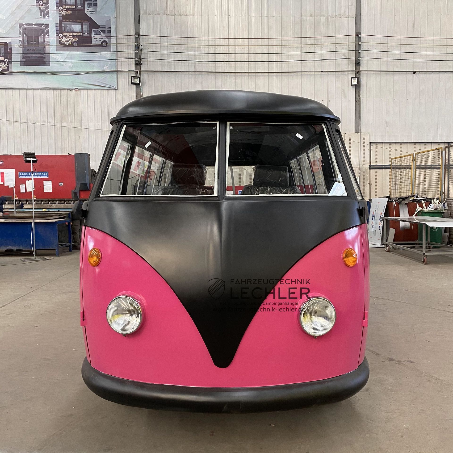 Ein rosafarbener und schwarzer VW-Bus mit markanten Scheinwerfern steht in einer Werkstatt.