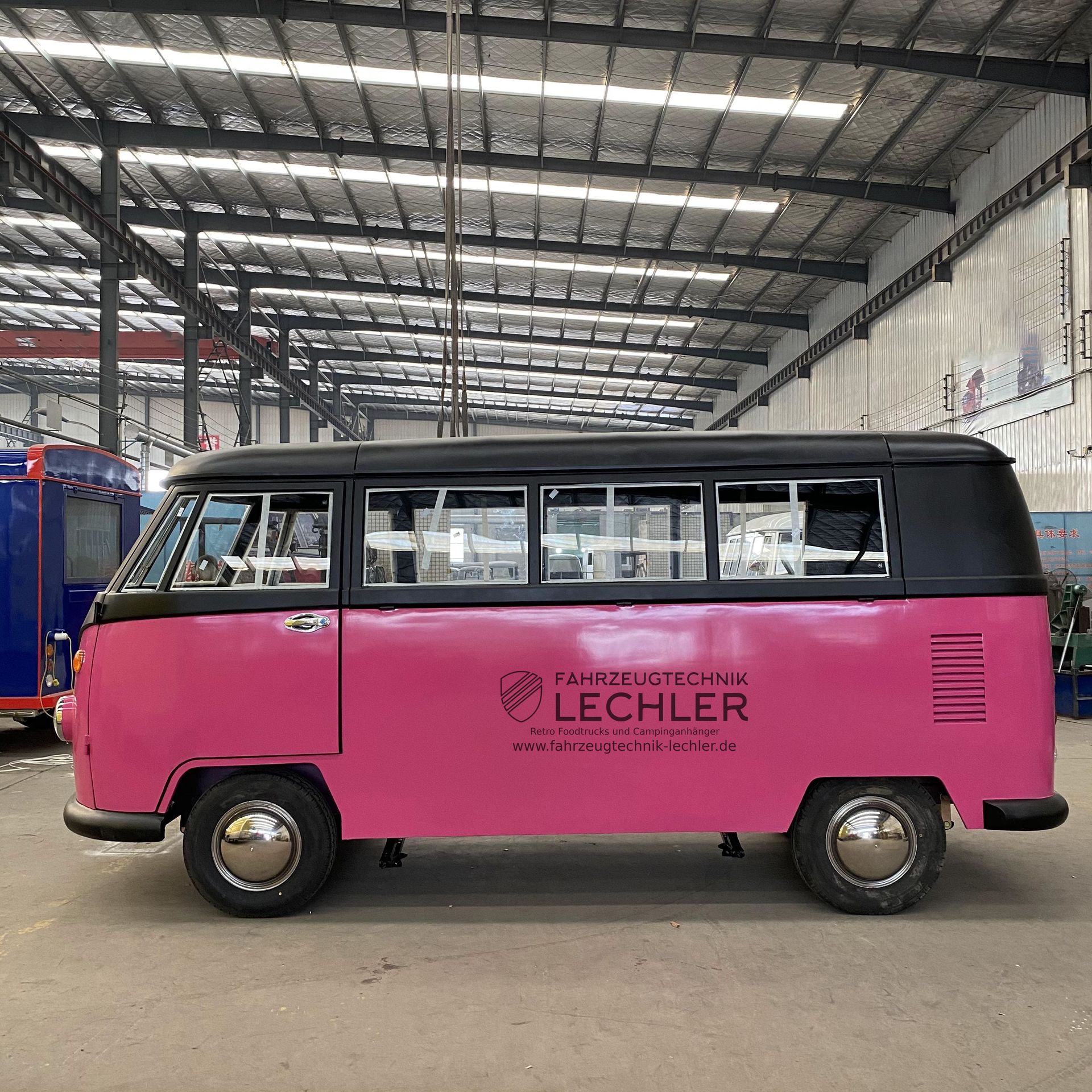 Ein rosafarbener und schwarzer Volkswagen-Bus steht in einer Metallhalle; an der Seite befindet sich das Firmenlogo.