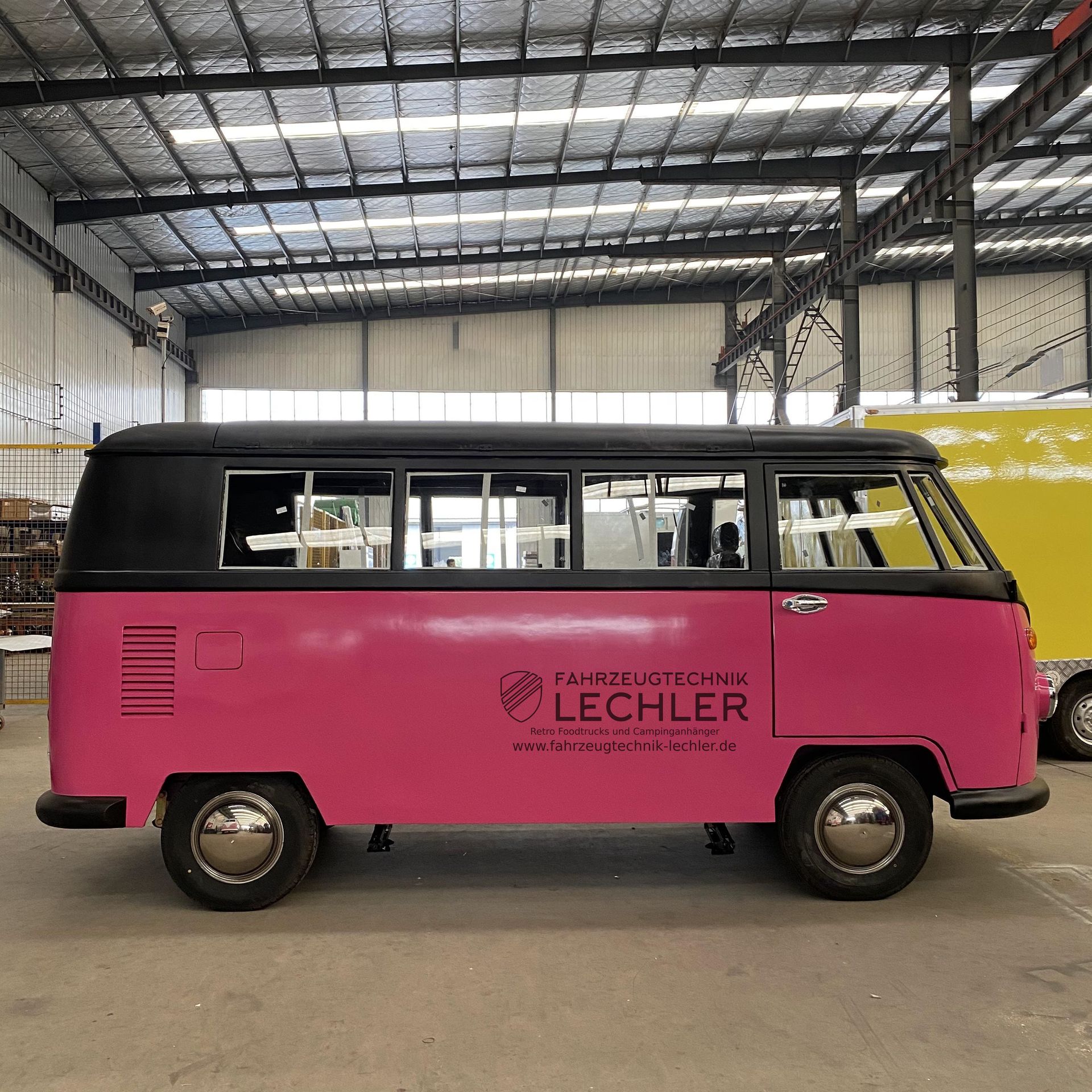 Ein rosafarbener und schwarzer VW-Oldtimerbus in einer Lagerhalle; an der Seite befindet sich das Logo von
