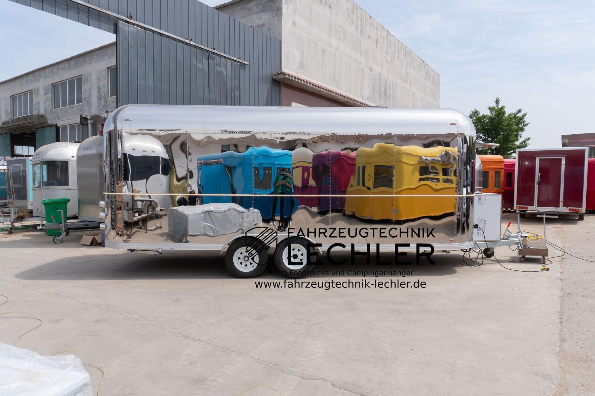 Ein glänzender, silberner Imbisswagen mit weißen Rädern, der die farbenfrohen Strukturen draußen auf einer gepflasterten Fläche reflektiert.