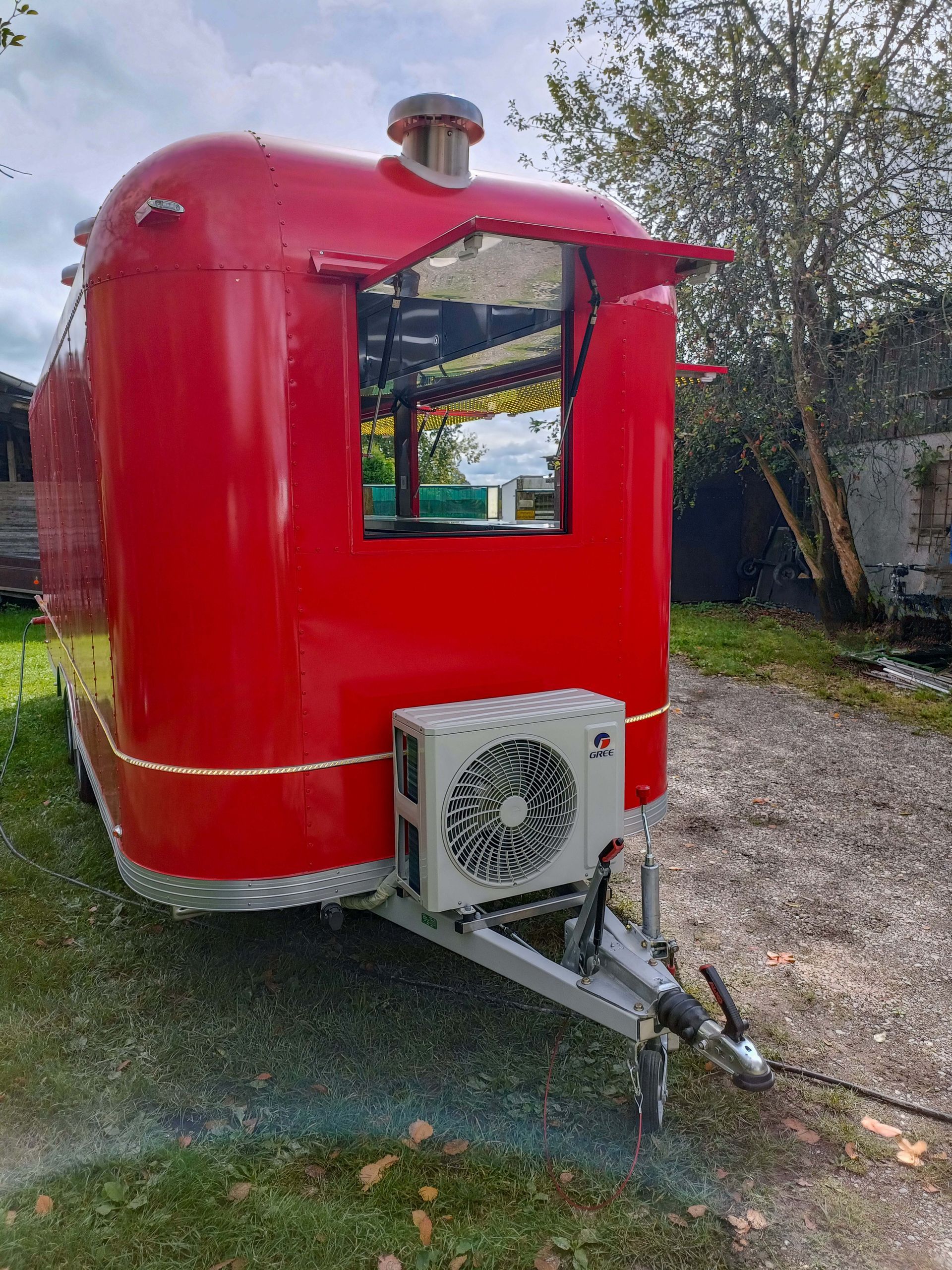 Roter Imbisswagen mit offenem Fenster, kleiner Markise und Klimaanlage, geparkt auf einer Rasenfläche.