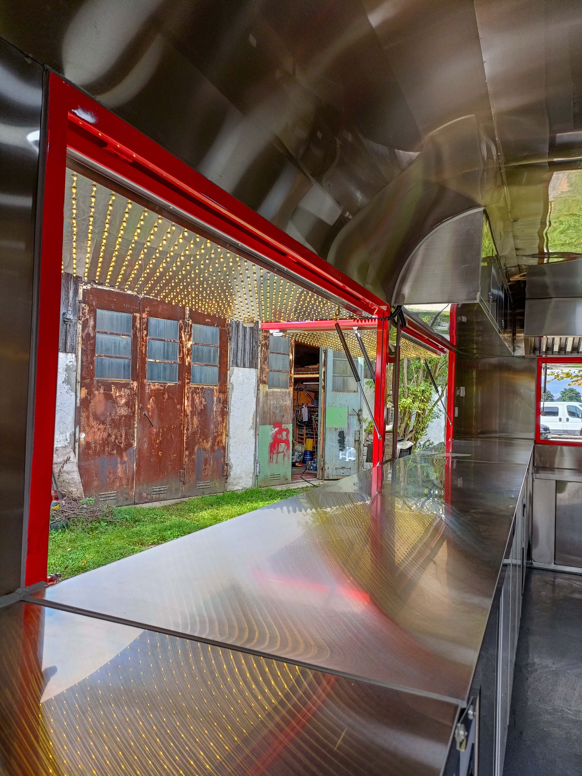 Innenansicht eines Imbisswagens mit rot umrandetem Ausgabefenster, das den Blick auf ein verwittertes Gebäude freigibt. Glänzende Metallarbeitsplatten.