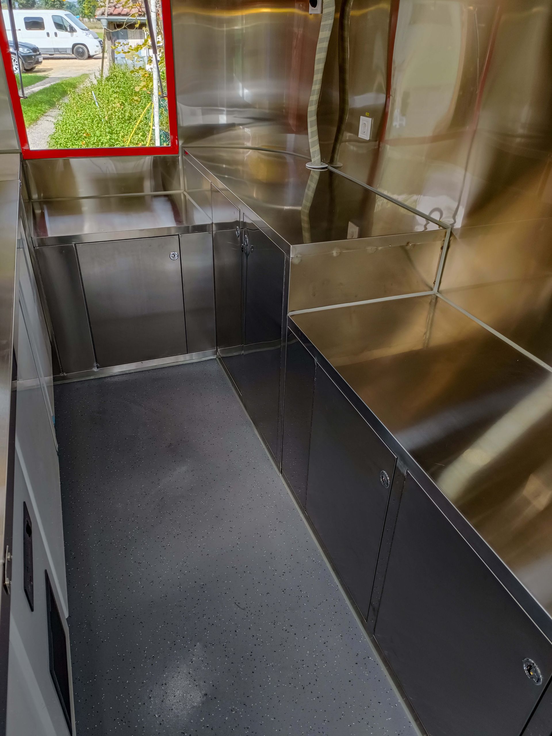 Innenraum eines Lieferwagens mit unfertigen Metallwänden und Regalen, links ein Fenster mit rotem Rahmen.