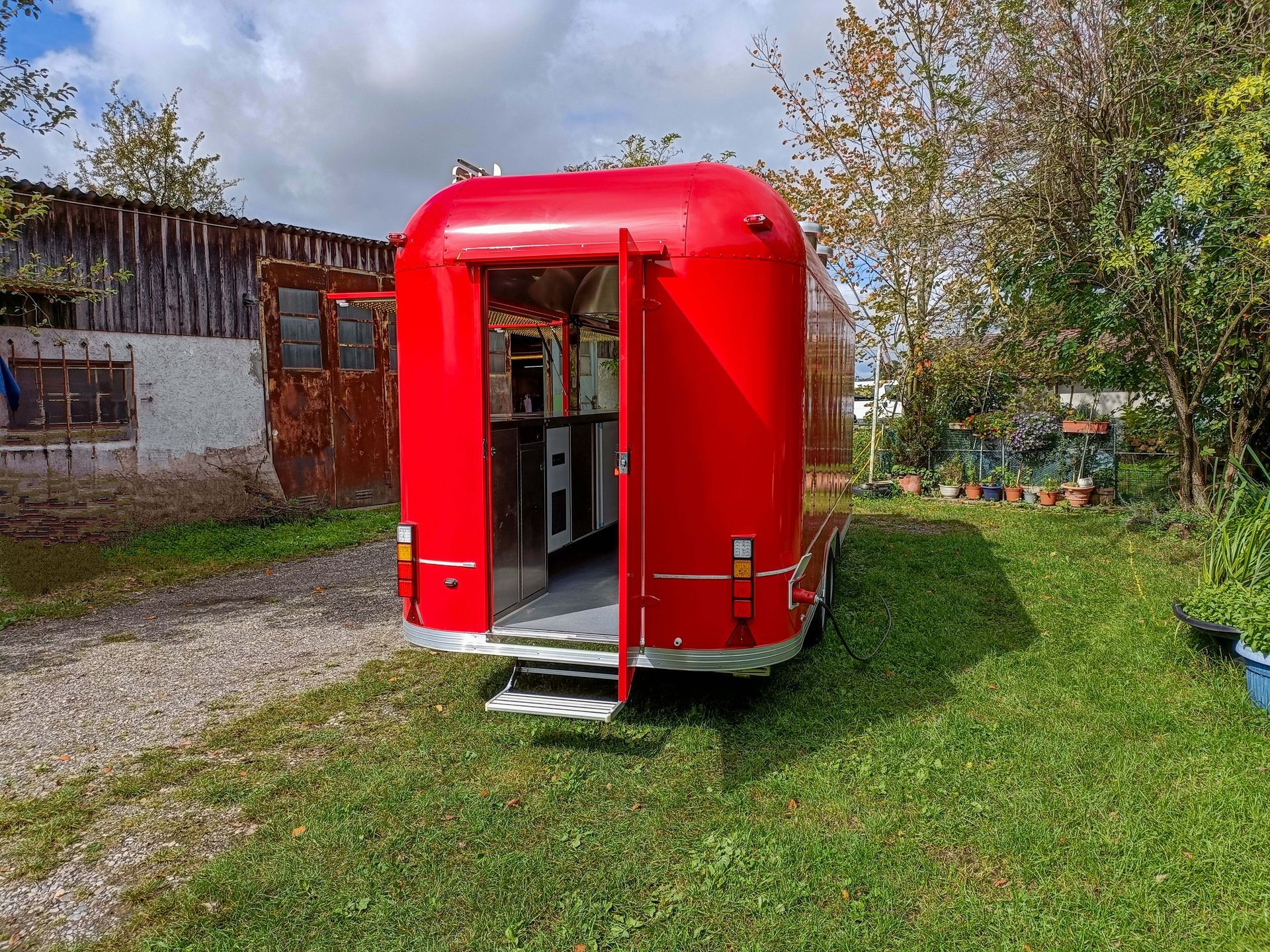 Roter Imbisswagen mit offenen Türen, auf einer Rasenfläche geparkt. Im Hintergrund Gebäude und Bäume.