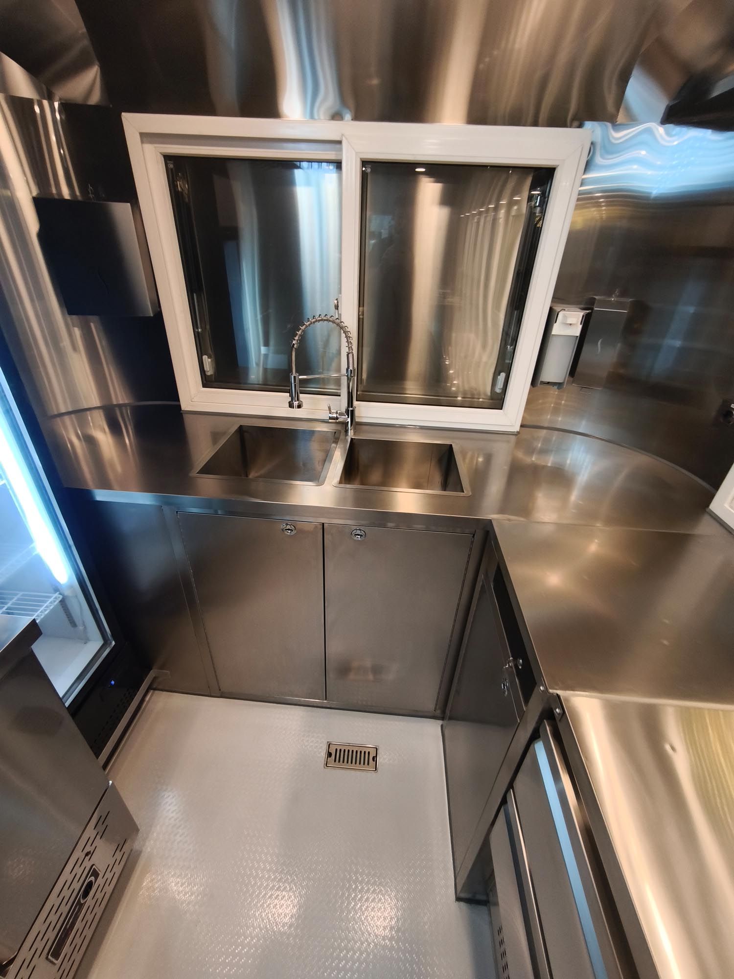 Innenansicht eines Imbisswagens mit Edelstahlflächen, Spüle, Schränken, Fenster und weißem Boden.
