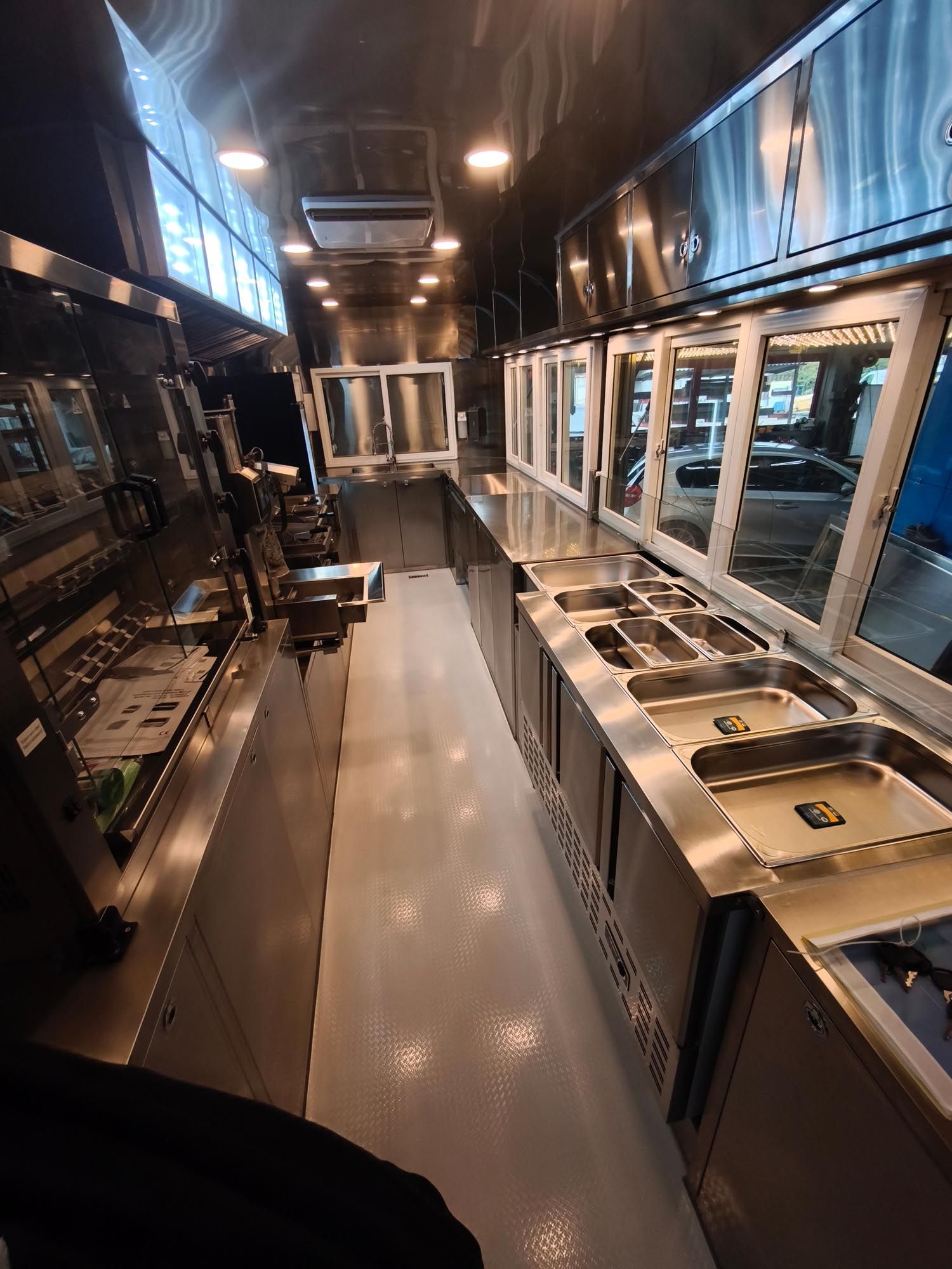 Innenansicht eines leeren Imbisswagens, Küchengeräte aus Edelstahl, weißer Boden, Glasfenster.