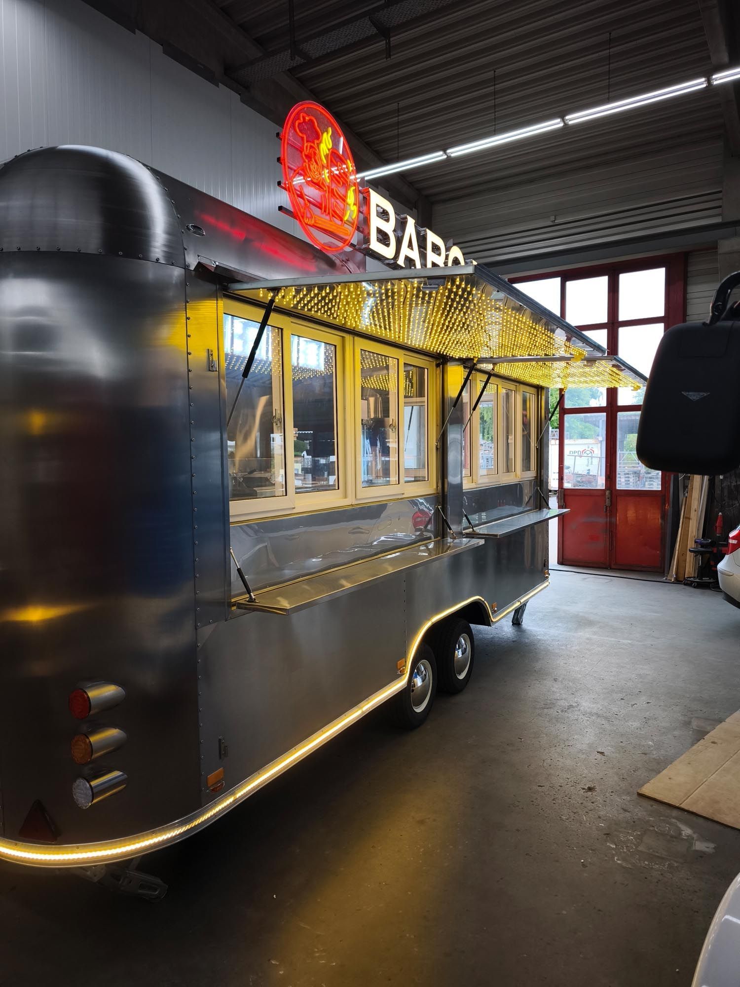 Ein glänzend silberner Imbisswagen mit dem Schild „BAR“, Markise und beleuchteten Kanten in einer Garage.