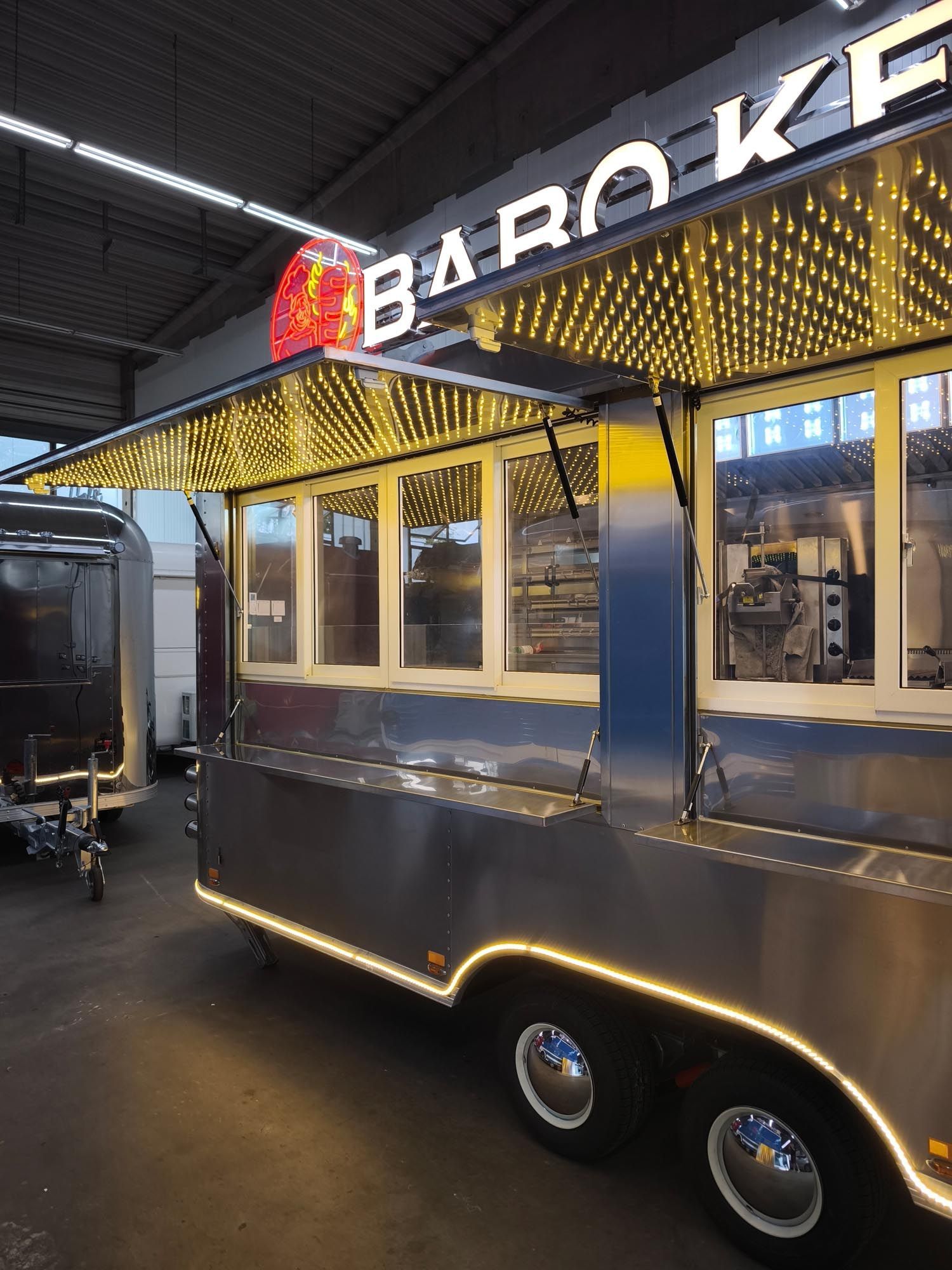 Imbisswagen mit beleuchtetem „BARO KEB“-Schild. Silberne Außenlackierung, Markise mit Beleuchtung, Fenster.
