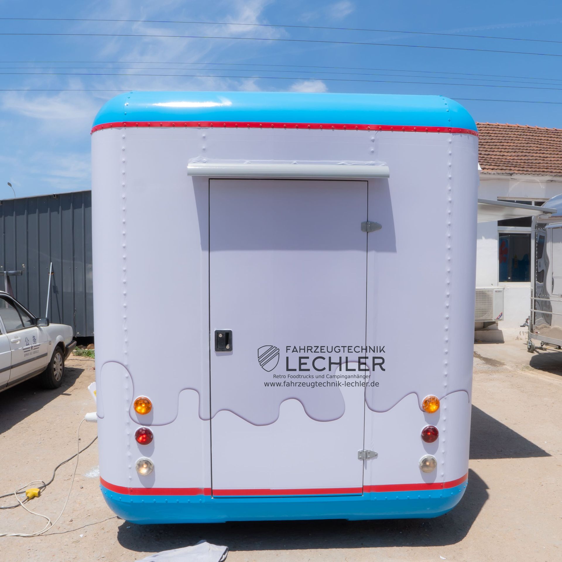 Rückansicht eines weißen Imbisswagens mit blauen und roten Akzenten, Tür und Logo; im Freien geparkt.