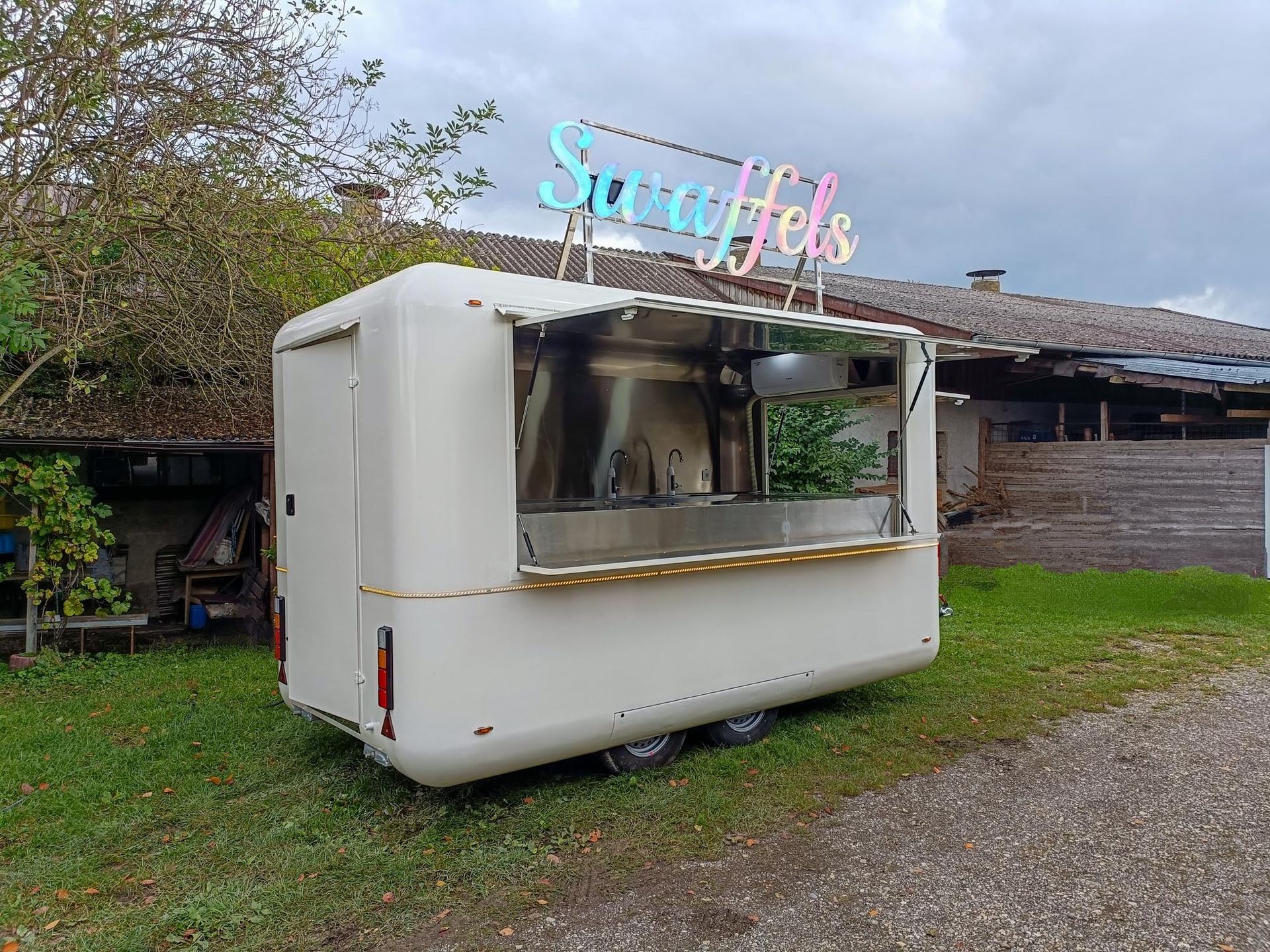 Weißer Imbisswagen mit offenem Ausgabefenster und dem Schild „Spuffels“ auf dem Dach. Steht auf einer Rasenfläche.