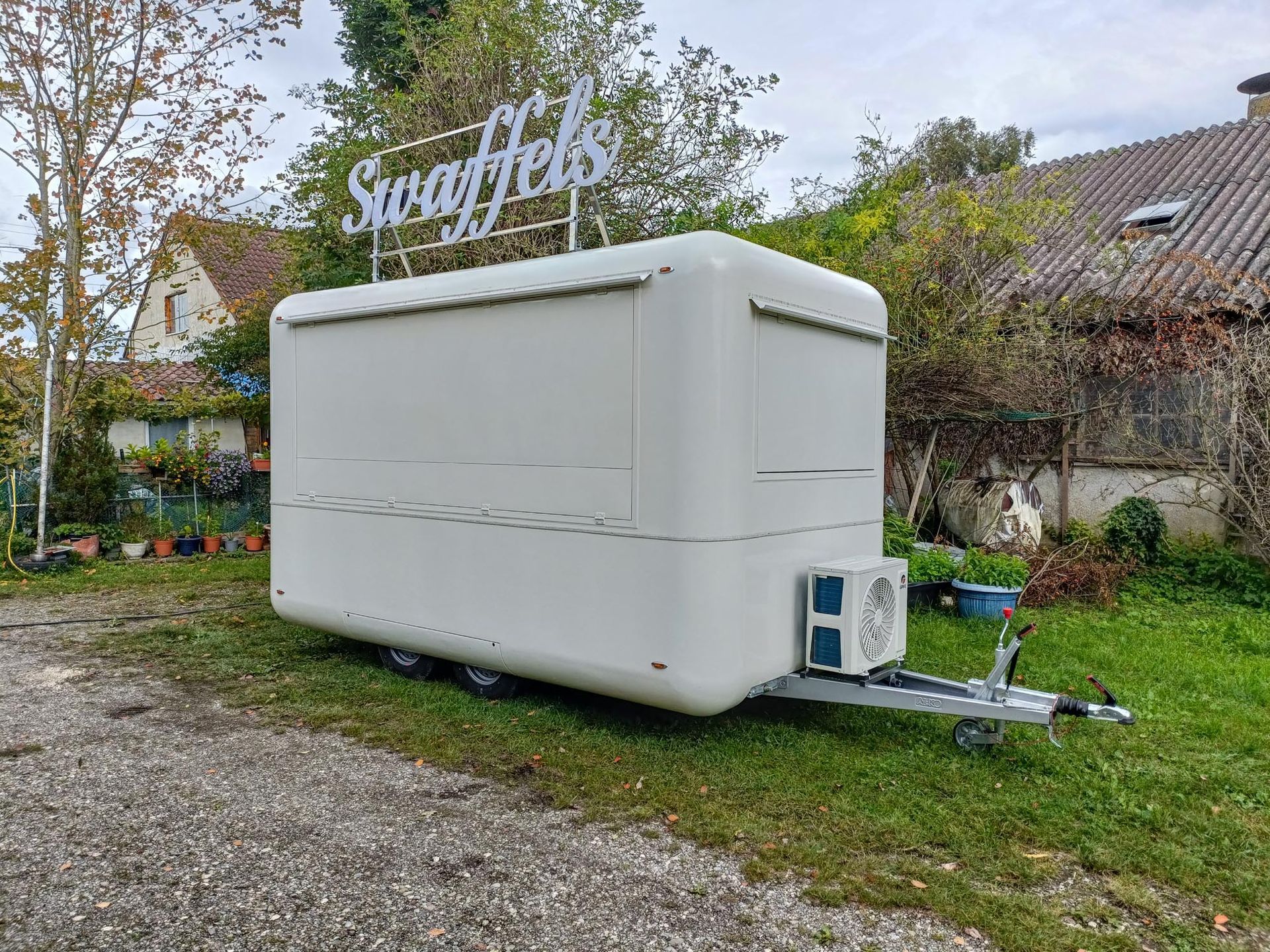 Weißer Imbisswagen mit dem Schild „Swaffels“ auf dem Dach, geparkt auf einer Rasenfläche. Im Hintergrund Bäume und Gebäude.