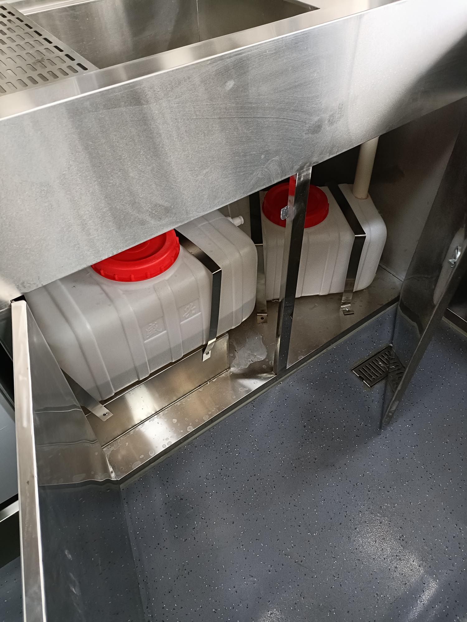 Zwei rechteckige Wassertanks mit roten Deckeln und schwarzen Bändern unter einem Edelstahlwaschbecken.