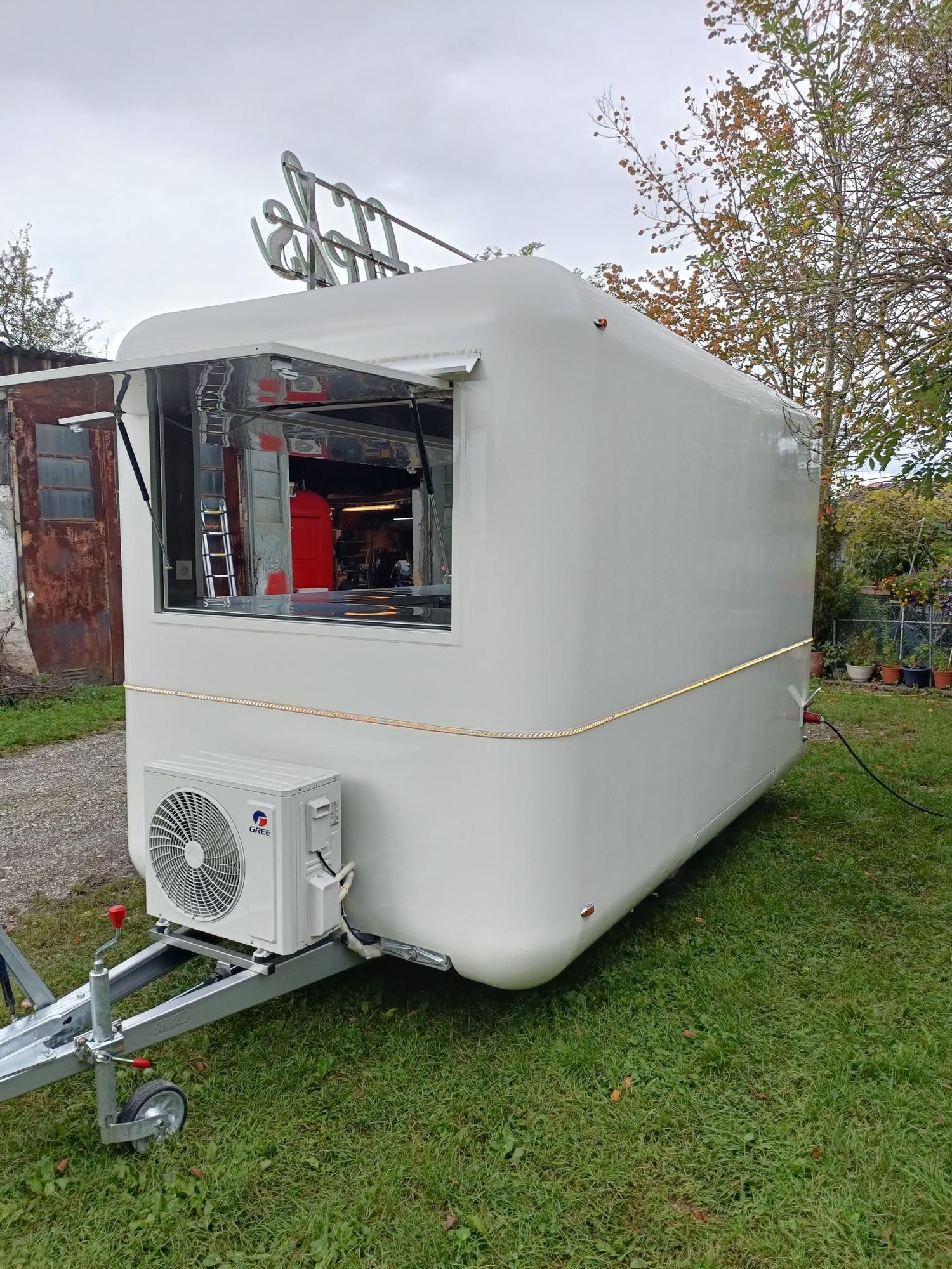 Weißer Imbisswagen mit Ausgabefenster an einer Anhängerkupplung, geparkt auf einer Rasenfläche.