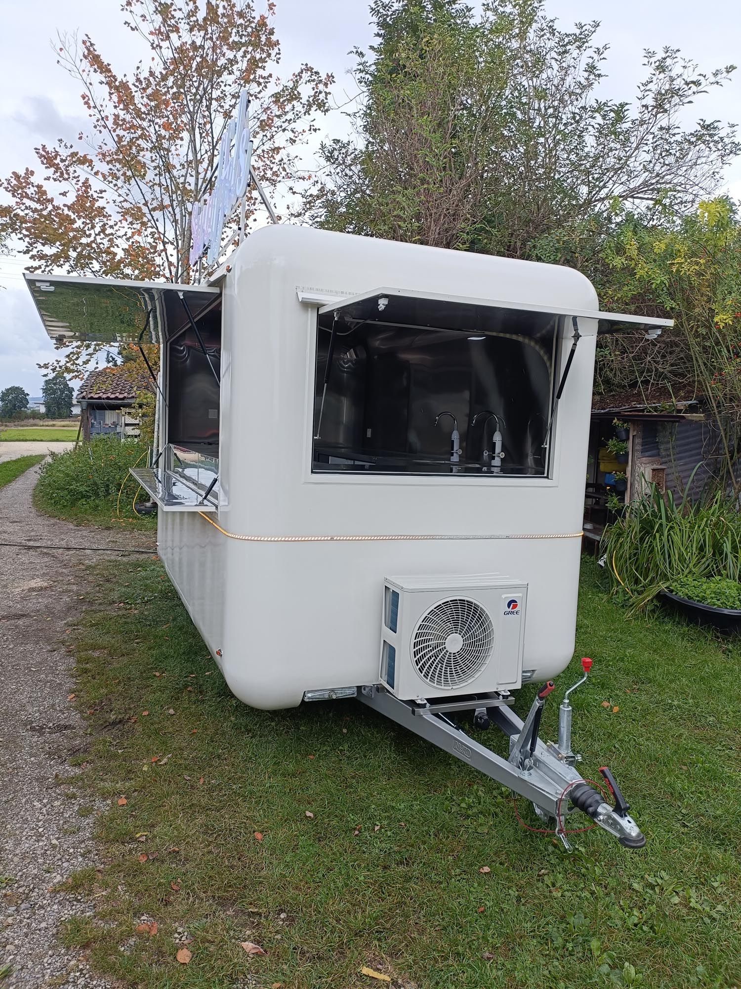 Weißer Imbisswagen auf einer Rasenfläche, mit offenen Ausgabefenstern und Klimaanlage.