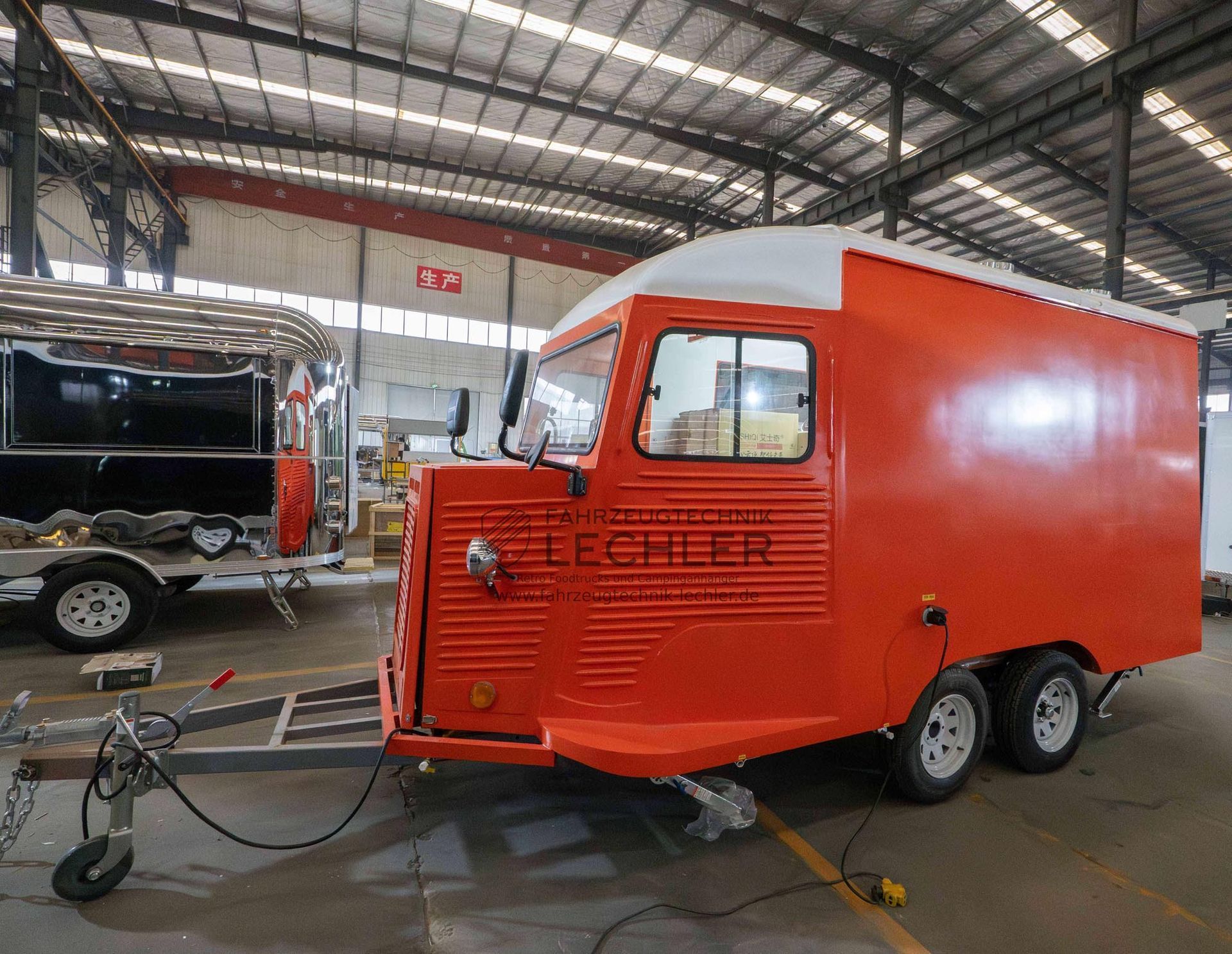 Ein orangefarbener Imbisswagenanhänger mit weißem Dach, weißen Rädern und Anhängerkupplung steht in einem großen Gebäude.