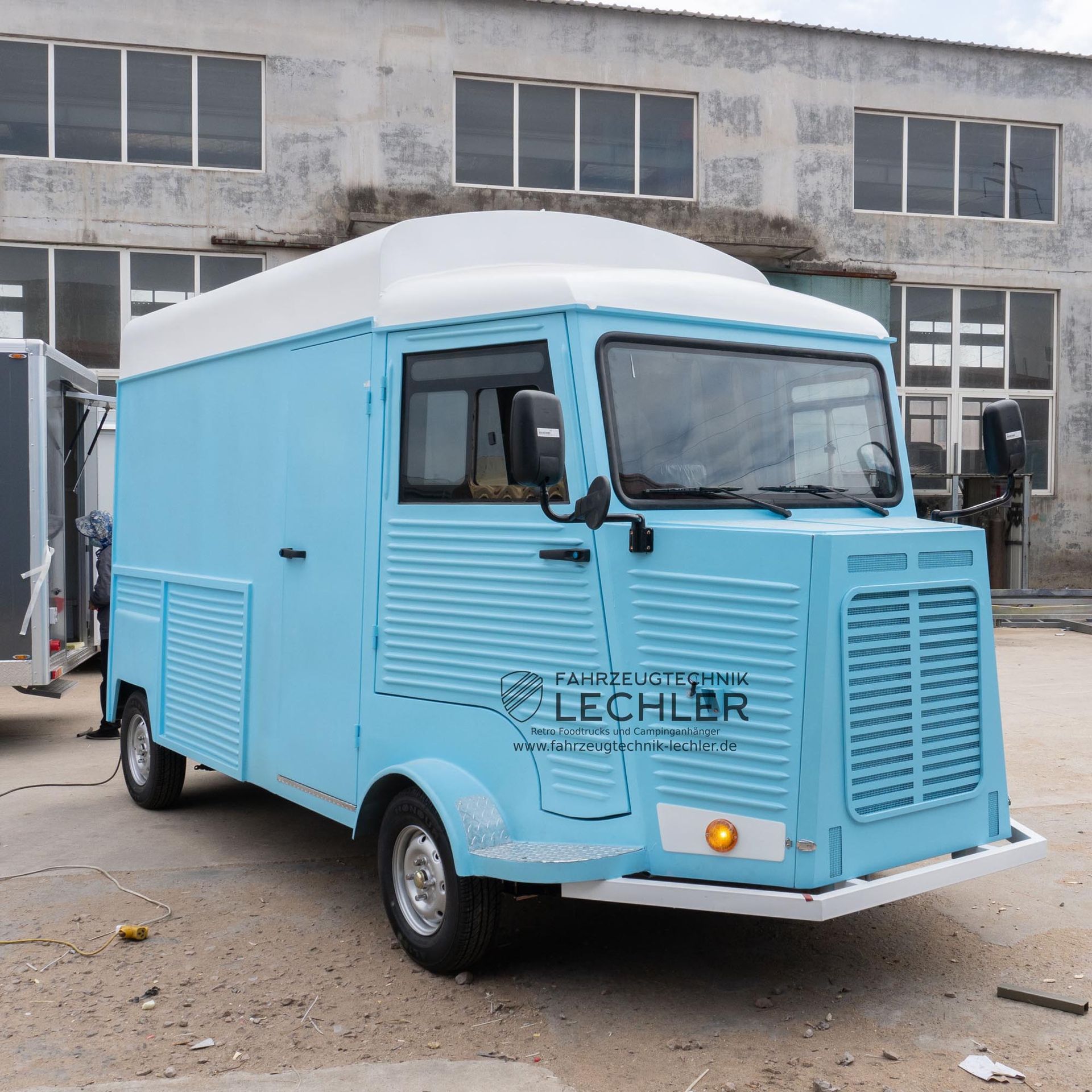 Hellblauer Imbisswagen mit weißem Dach, der vor einem Gebäude parkt.
