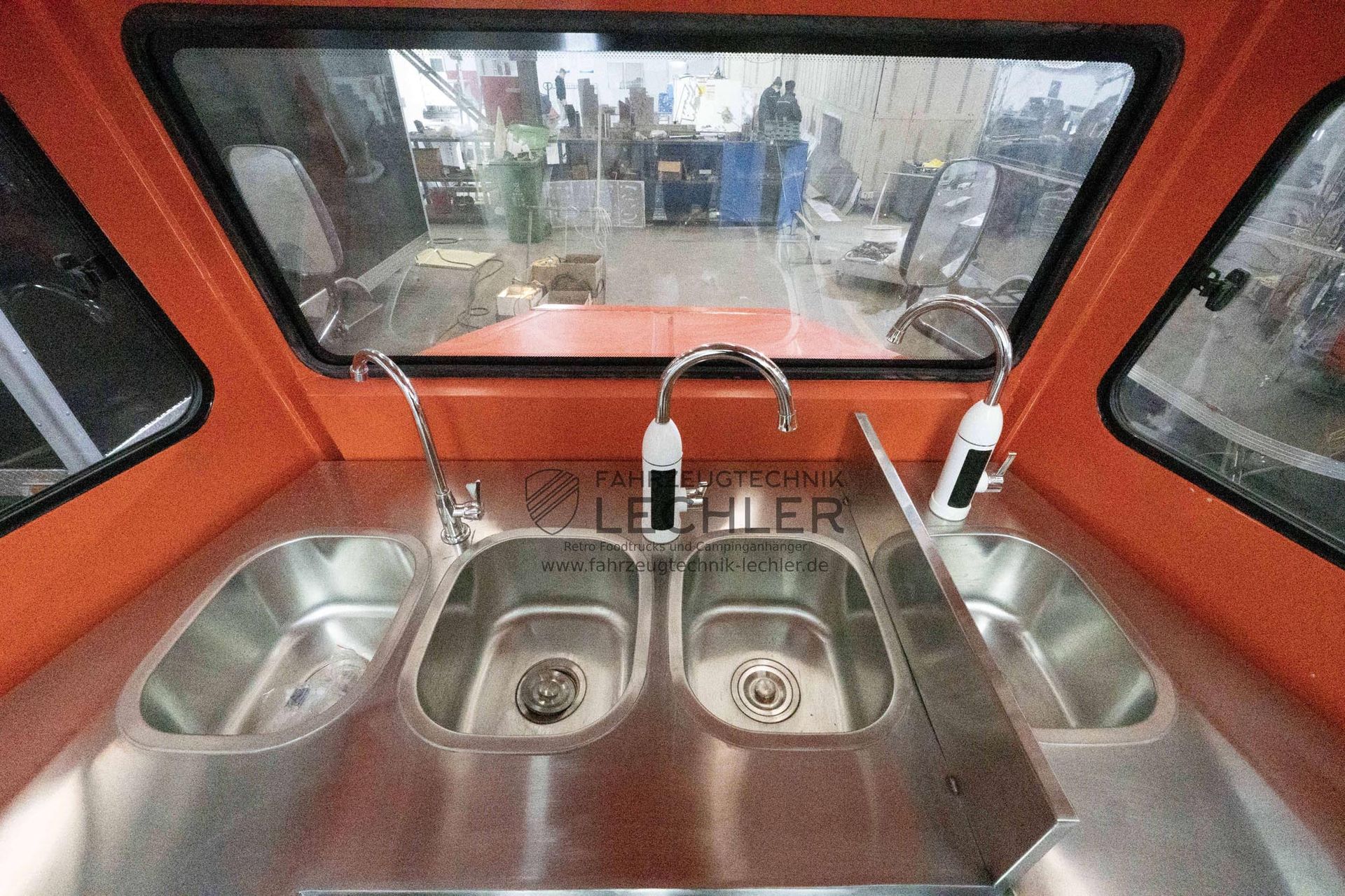 Im Inneren eines Fahrzeugs: Drei Edelstahlspülen mit Armaturen sind in eine orangefarbene Arbeitsplatte eingelassen. Ein Fenster gibt den Blick auf ein Geschäft frei.