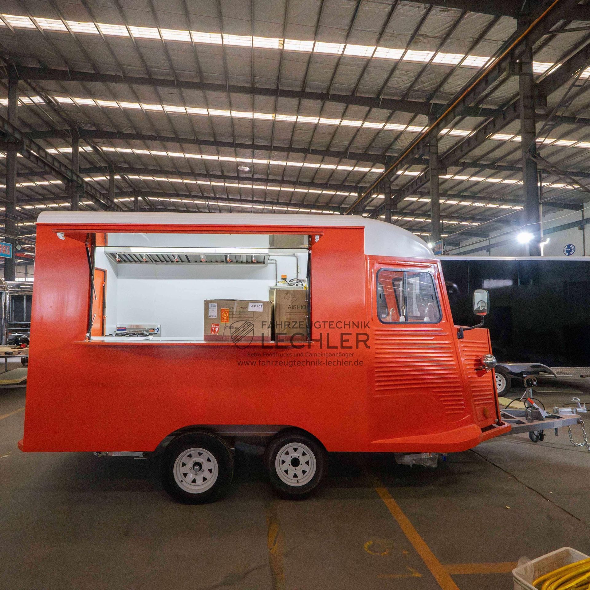 Ein orangefarbener Imbisswagen mit weißen Zierelementen und einem Ausgabefenster steht in einer großen Lagerhalle.