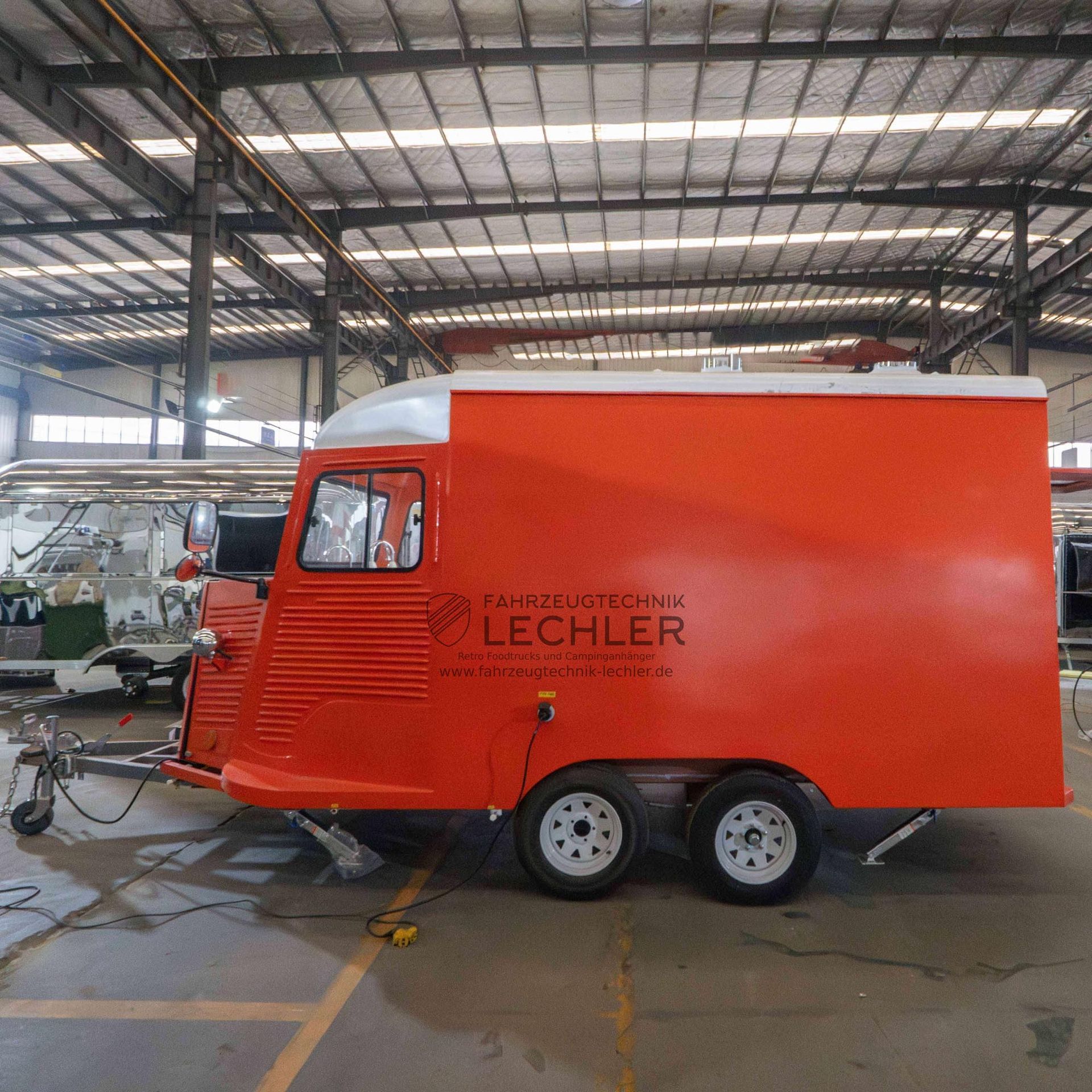 Oranger Imbisswagen mit weißem Dach, Logo an der Seite, zwei Rädern, im Innenbereich geparkt.